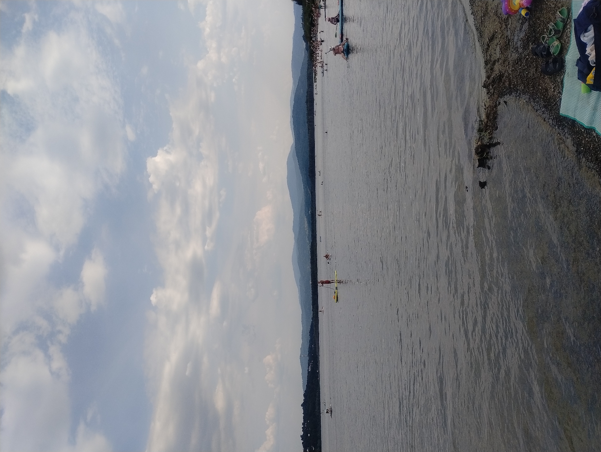The image depicts a wide, calm lake with a shoreline in the foreground and a hilly, forested landscape in the distance. The sky is partly cloudy with scattered white clouds against a pale blue background. Several people are visible on the lake, engaged in recreational activities.
In the middle ground, numerous individuals are on paddleboards. One person in a red top is standing upright on a yellow paddleboard, holding a paddle. Another person on a blue paddleboard is also visible. In the background, there are more paddleboarders and swimmers in the water.
The shoreline in the foreground is covered with dark sand, pebbles, and some vegetation. On the shore, there is a striped mat with a bag containing clothing, and a pair of black sandals and green sandals. A colorful toy, possibly a ball, is also present.
The overall scene suggests a summer day at a recreational lake, likely in Zarzecze, Poland, given the context of the provided information.