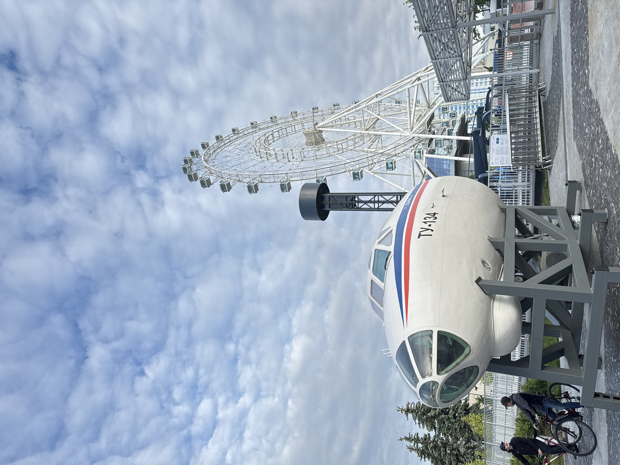 This outdoor image, likely taken at VDNKh in Moscow, Russia, features the cockpit and front fuselage of a Tupolev Tu-134 aircraft on display, set against a backdrop dominated by a large white Ferris wheel.
The Tupolev Tu-134 aircraft section, painted white with distinctive red and blue stripes running along its side, is mounted on a robust grey metal frame. The Cyrillic text "ТУ-134" is clearly visible on its exterior. Below the aircraft and towards the right foreground, two individuals, presumably men, are present with bicycles. One person stands next to a red bicycle, while the other is close to a black bicycle.
In the background, the prominent structure of a large white Ferris wheel stands tall against a cloudy, bright daytime sky. Its intricate spoke design and numerous enclosed passenger capsules are visible. Sparse green trees and architectural elements of distant buildings can be seen beyond the Ferris wheel and the display area. Metal railings and paved ground further define the public setting, suggesting an exhibition or amusement park environment. The scene depicts a calm daytime setting with no discernible specific event in progress beyond the display of the aircraft and the presence of park visitors.