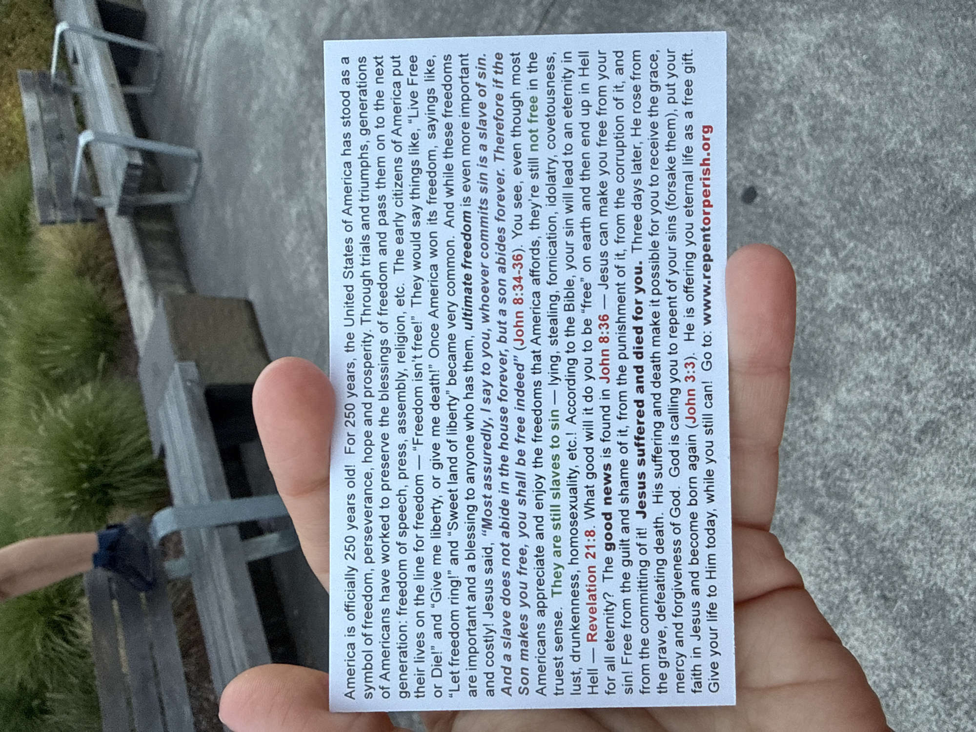 A close-up outdoor shot shows a person's thumb and forefinger holding a small white card with text printed on it. The card discusses freedom, sin, and salvation, referencing biblical scripture. Behind the card and hand, a blurred background reveals a park-like setting with a wooden bench, concrete structures, and greenery. The lighting suggests it is daytime.