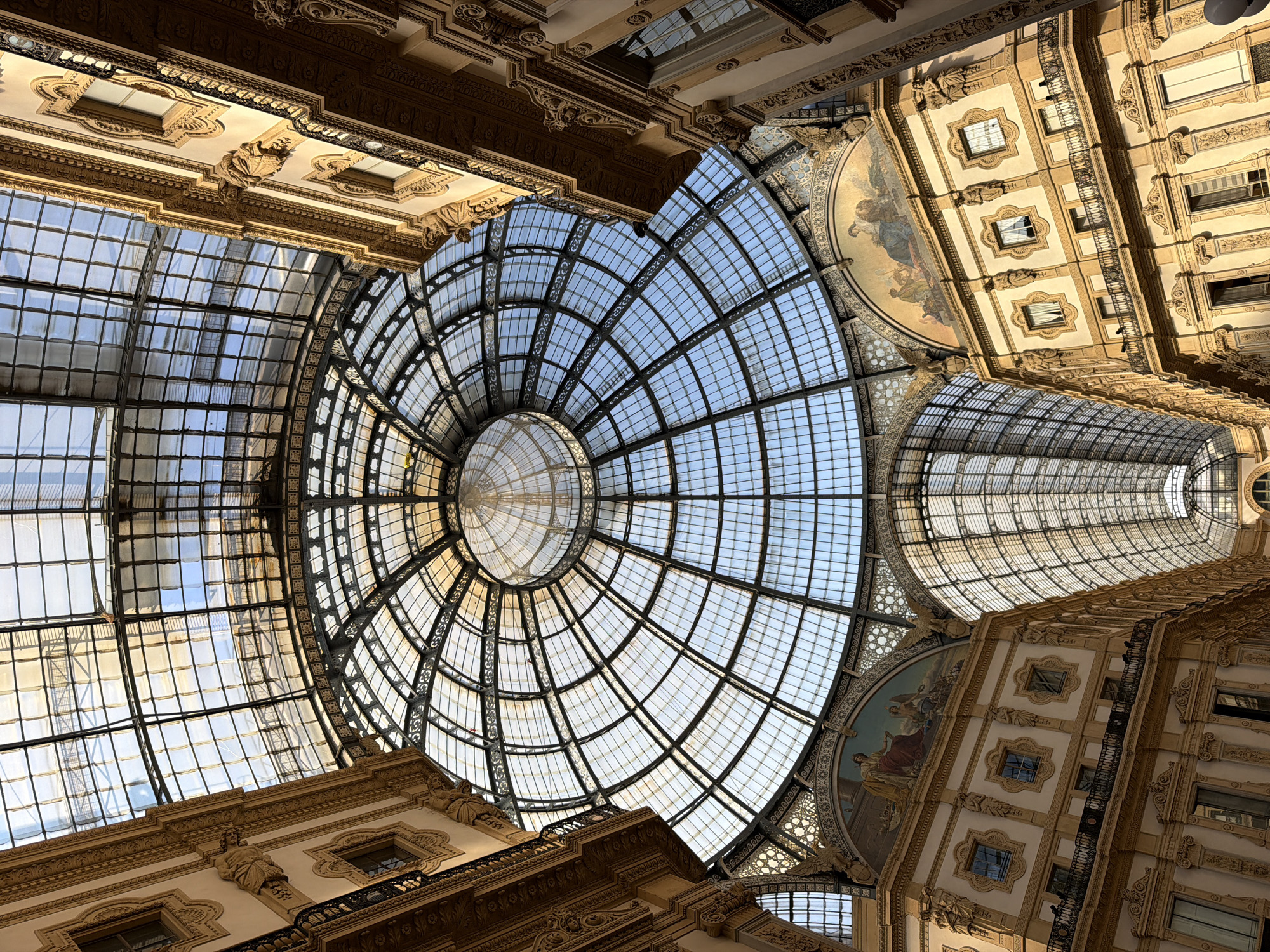 The image captures an upward view of the Galleria Vittorio Emanuele II in Milan, Italy. The scene showcases the architectural grandeur of the structure, focusing on the glass and iron dome and the surrounding buildings. The dome's intricate framework is visible against the daylight, highlighting its geometric design. The surrounding buildings feature ornate facades with decorative elements, windows, and architectural details. No people are visible in the image. The photograph emphasizes the architectural beauty and scale of the Galleria Vittorio Emanuele II.