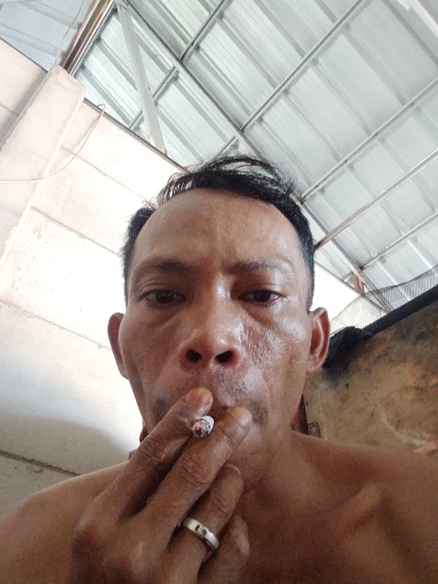 A close-up, low-angle shot depicts a shirtless, middle-aged Asian man with short, dark hair, fair skin, and visible pores smoking a cigarette. A silver ring is visible on his left ring finger. The background consists of corrugated metal roofing and structural beams typical of a building in Bukit Ketok, Indonesia. A blurry, nondescript object sits partially out of focus to the right.