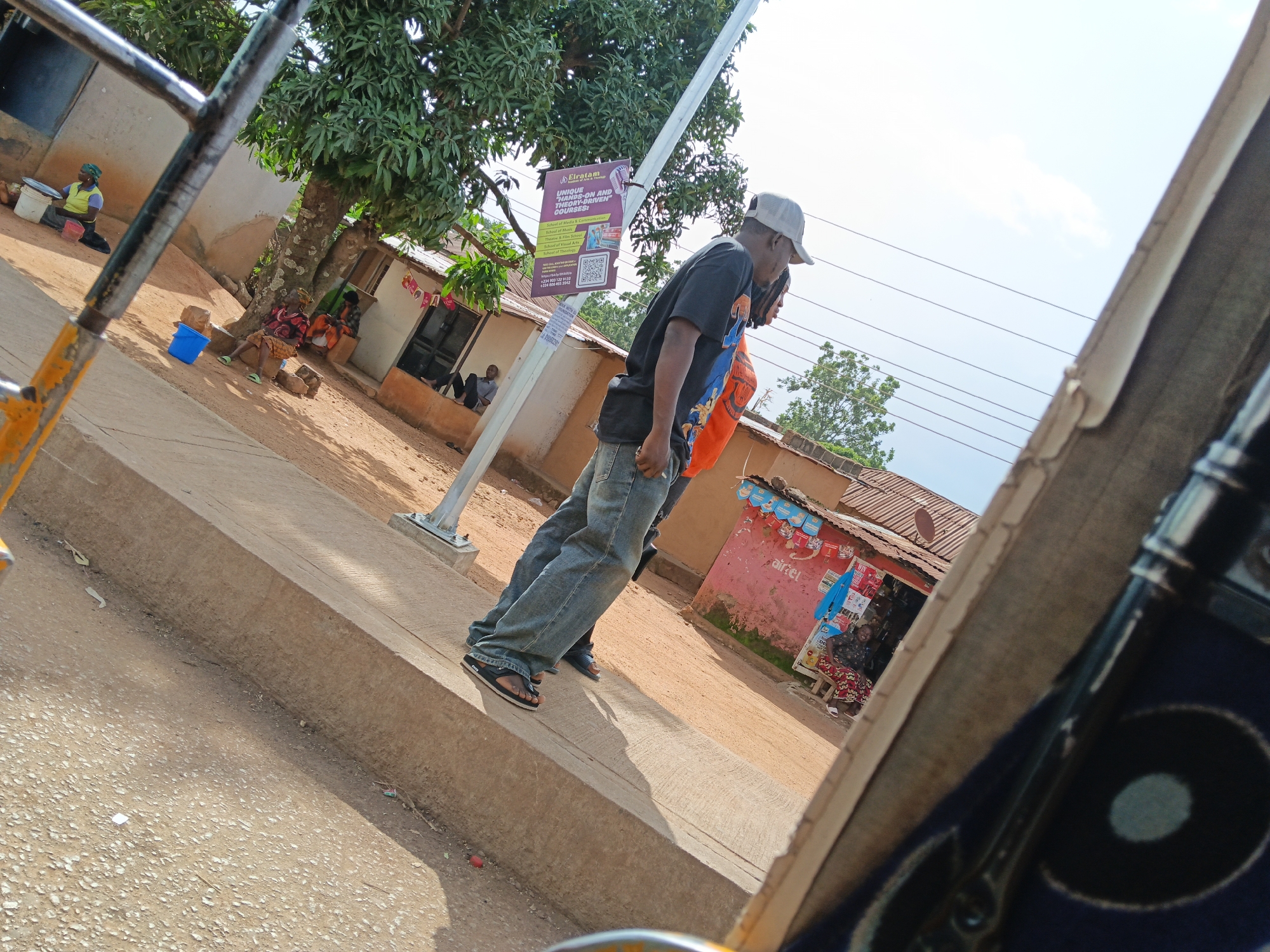 A man wearing a baseball cap, a dark t-shirt, and blue jeans stands on a concrete curb. He is facing to the right and looking down. Behind him, another person wears an orange shirt. In the background, several people are seated outside a building with a red wall, and a woman in a yellow vest is visible near some buckets. There are trees and a utility pole in the scene. The ground is dirt. The overall setting appears to be an outdoor marketplace or street.