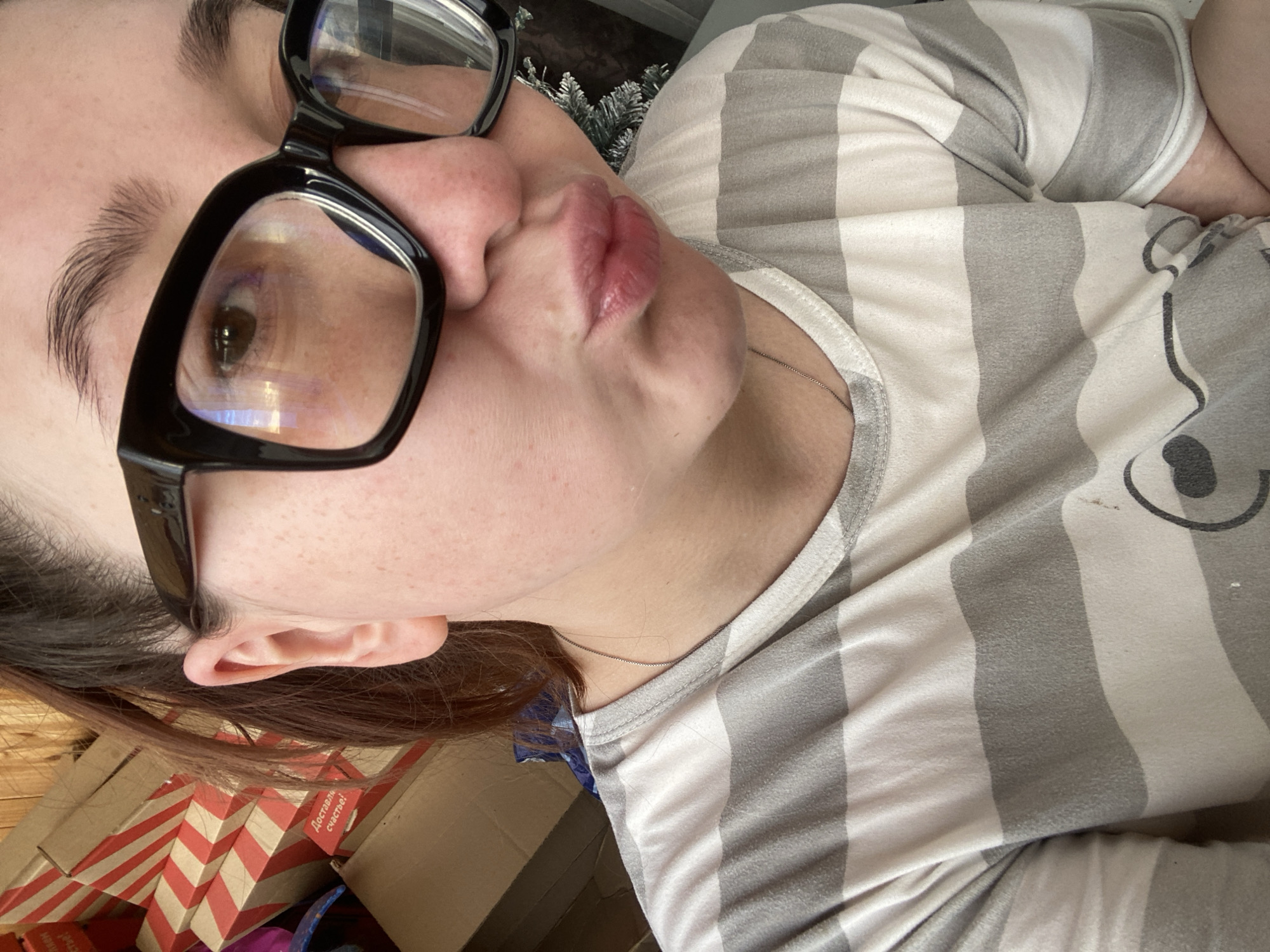 A close-up, angled selfie shows a person wearing black-framed eyeglasses and a gray and white striped T-shirt. The person is puckering their lips. The background includes a cardboard box with red and white stripes and Russian text, and a hint of artificial green foliage. The image location cannot be determined as the city of Chaikovsky, Russia based on the available information.