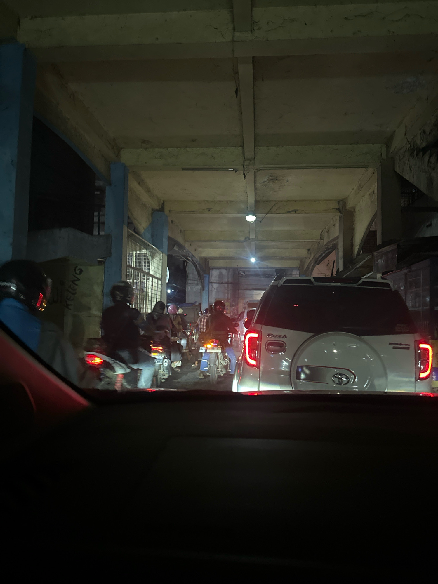 The image captures a traffic jam at night, possibly on a road with overhead structures in the city of Binjai, Indonesia. The perspective is from inside a vehicle, looking forward. In the foreground, the dark interior of the car is visible. The scene ahead is dominated by various vehicles, primarily motorcycles with riders wearing helmets, and a white SUV with its taillights illuminated. Some of the motorcycles are in motion, while others are stationary in the traffic.
The environment appears to be an urban setting, with concrete structures forming an overhead passage or bridge. The lighting is dim, characteristic of nighttime, with streetlights or vehicle lights providing illumination. A yellow wall with blue lettering is visible on the left side, adding to the urban context. The overall impression is one of a congested road at night in a densely populated area.