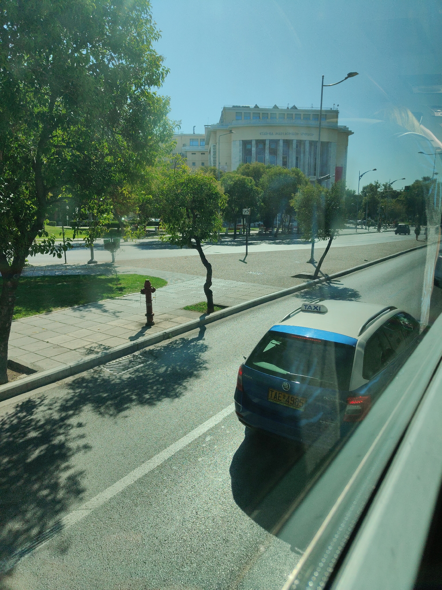 The image depicts a street scene in Thessaloniki, Greece, with a blue Skoda Octavia taxi driving on the right side of the road. The taxi has a "TAXI" sign on its roof and a license plate reading "TAE 4985". In the background, a large, light-colored building with columns and arched windows is visible, likely a public institution or historical landmark. Several trees line the street, casting shadows on the sidewalk and road. A red fire hydrant is present on the sidewalk. The sky is clear and blue, indicating a sunny day. The image appears to be taken from inside a vehicle, with reflections visible on the window.