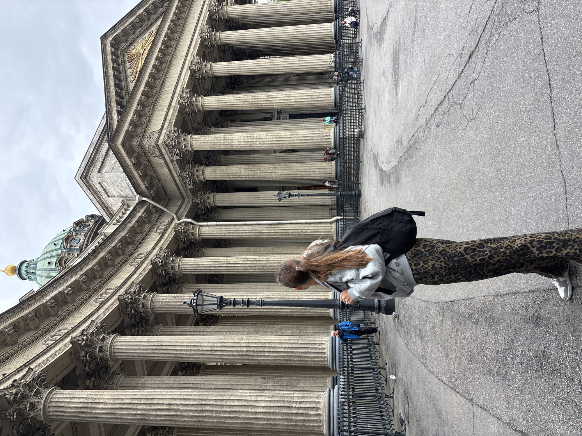 The image depicts a woman standing on a paved surface in front of a large, ornate building. The building is recognizable by its many Corinthian columns, topped by elaborate entablature and a prominent dome in the background, consistent with the Kazan Cathedral in Saint Petersburg, Russia. The woman has long brown hair and is wearing a light gray jacket, leopard-print pants, and white sneakers. She carries a black backpack. She appears to be looking down at something near a black metal fence and a lamppost. Other people are visible in the background, standing on the building's stairs and near the columns. The sky is overcast.