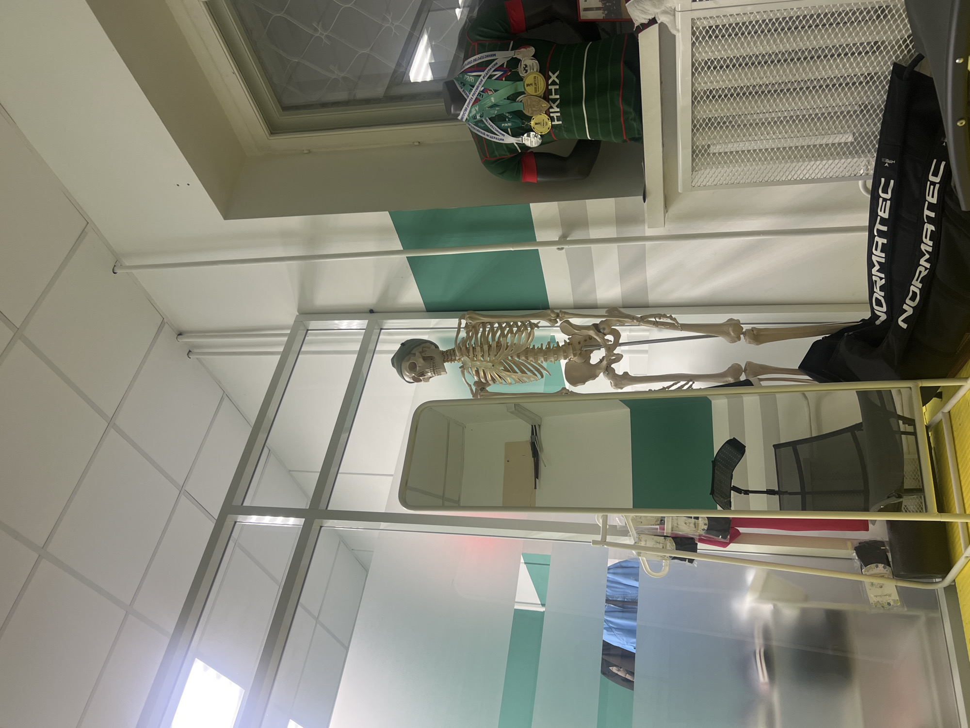 This media file presents an indoor scene, likely within a sports medicine facility, clinic, or an educational setting in Kazan, Russia. The room is brightly lit and features modern design elements, including transparent glass partitions and a white ceiling composed of square panels.
A life-size anatomical skeleton model stands prominently in the center-left of the image, positioned behind a white, possibly metallic, framed structure that resembles a bed or examination table. To its right, a mannequin is displayed, adorned in a dark green and red sports jersey featuring white horizontal stripes. The jersey clearly shows the Cyrillic text "HKHX" and "МИНИСТЕРСТВО СПОРТА РОССИИ" (Ministry of Sport of Russia), along with the partially visible sponsor name "PARI." Multiple gold and silver medals hang around the mannequin's neck, signifying athletic achievements. Above the mannequin, a window with a mesh screen provides an external view.
Further to the right, a black bag with the brand name "NORMATEC" printed in white is visible, indicating the presence of specialized recovery or compression therapy equipment. The background wall features a distinctive horizontal green stripe against a lighter colored wall. The reflective glass partitions catch reflections of the room, including glimpses of what appears to be a black mesh chair and other equipment. The scene is static, devoid of any ongoing activity, and suggests a display or a functional setup within a professional environment.