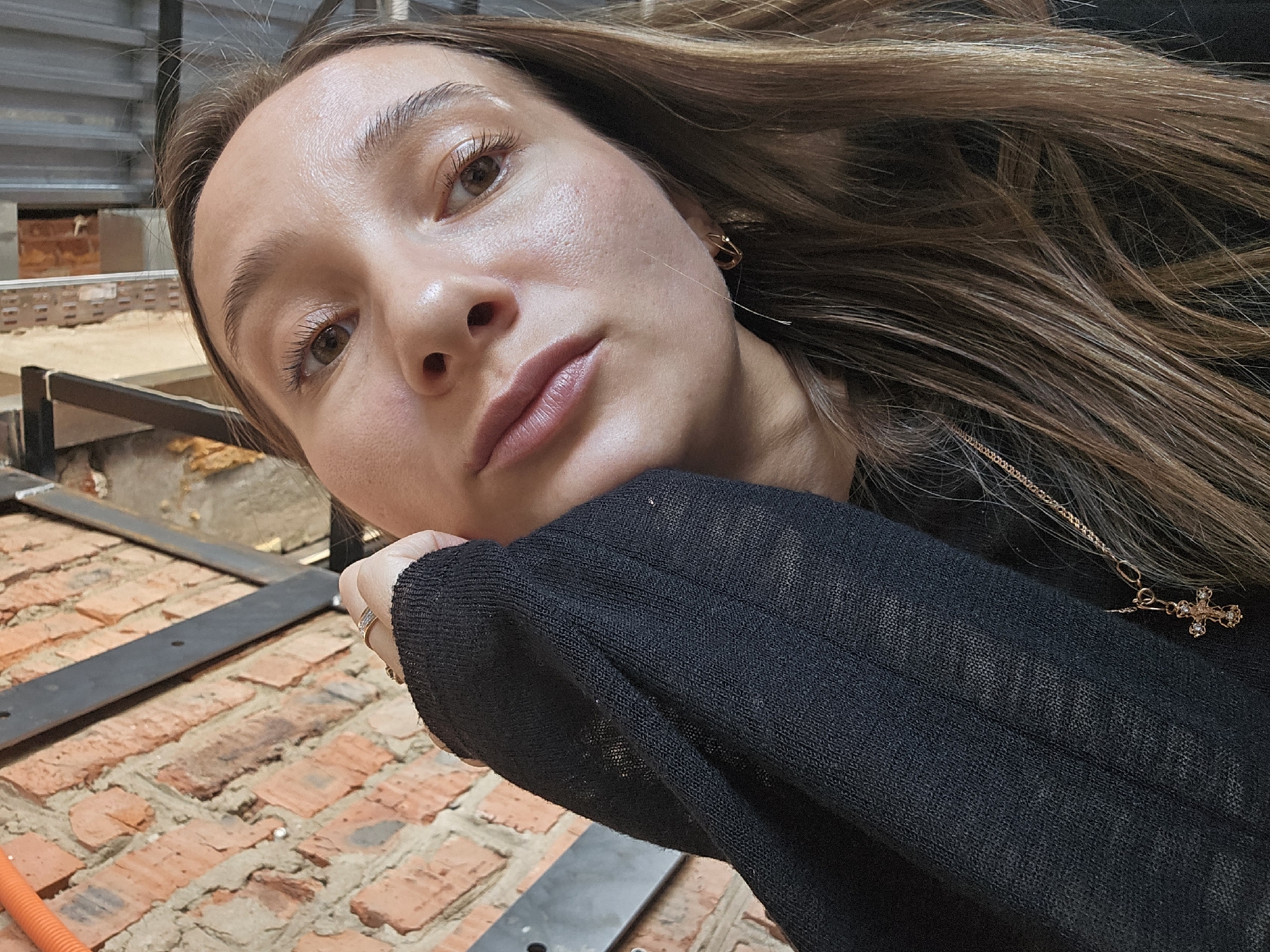 A close-up, slightly low-angle portrait captures a young adult female in an indoor setting within the city of Chaikovsky, Russia. The subject has medium-length light brown hair, brown eyes, and a neutral expression, looking directly at the camera. Her right hand, adorned with a gold-colored ring with small white stones on the ring finger, is positioned with the back of the hand resting against her right cheek and jawline. She wears a black, long-sleeved, fine-knit top. A thin gold chain necklace with a small, ornate, jewel-encrusted cross pendant is visible against her chest. A small gold hoop earring is present in her right ear. The background on the left features an exposed red brick wall with dark metal beams, suggesting an industrial or loft-style interior. The upper right background contains elements consistent with such an environment, including what appears to be corrugated metal paneling and other building materials.