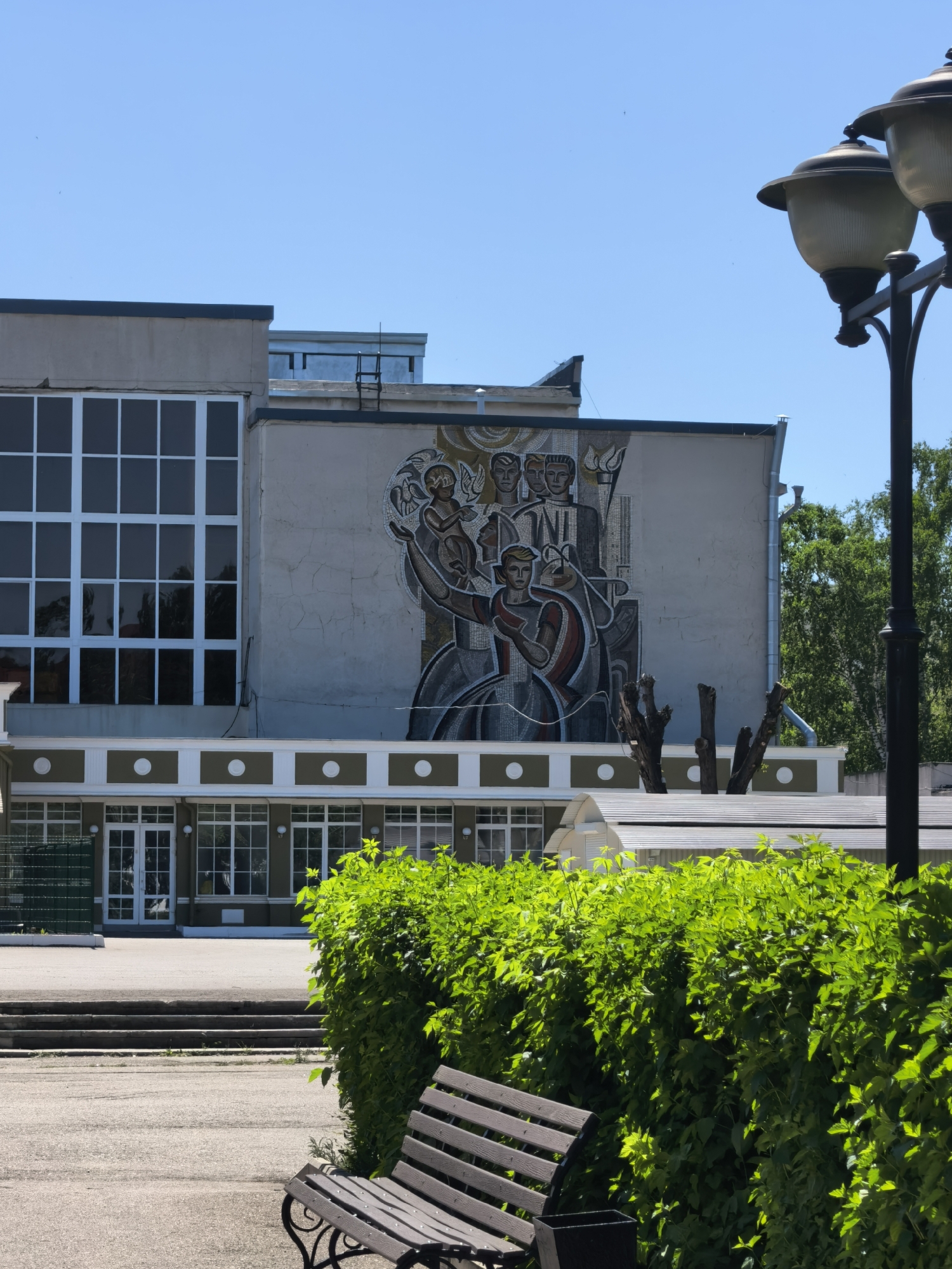 The image captures a building with a mosaic mural in Sterlitamak, Russia. The building has a concrete facade with a grid-patterned window section on the left and the mural on the right. A decorative row of circular and rectangular shapes runs along the lower edge of the upper building section. A lower level of the building is visible with multiple windows and doors.
A well-maintained hedge and a park bench are in the foreground. A vintage-style lamppost stands on the right side of the frame. The sky is clear and blue, indicating a sunny day. A trimmed tree is visible in front of the mosaic.