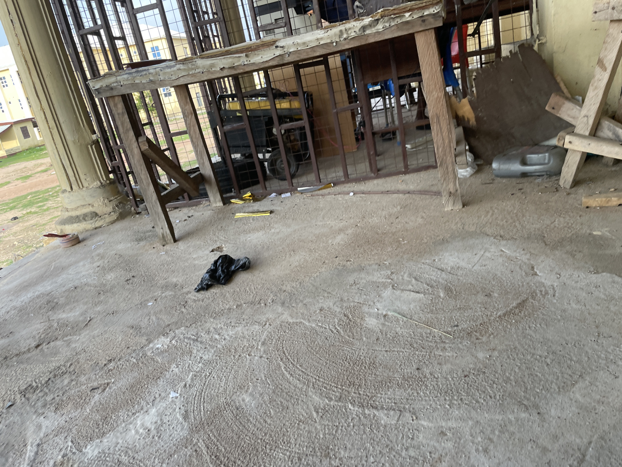The image depicts a rough, outdoor area in Jalingo, Nigeria, characterized by a concrete ground with visible textured patterns and debris. A black plastic bag is present on the ground, along with a metal object and some small white pieces that could be debris. To the right, a structure made of wood and metal grating is visible, appearing to house various objects. These include what looks like a generator, a cardboard box, a grey plastic container, and what could be a red bucket or container. The background reveals parts of buildings with windows, suggesting an urban or semi-urban environment. The overall scene suggests a storage or work area, possibly an outdoor section of a property or a yard.