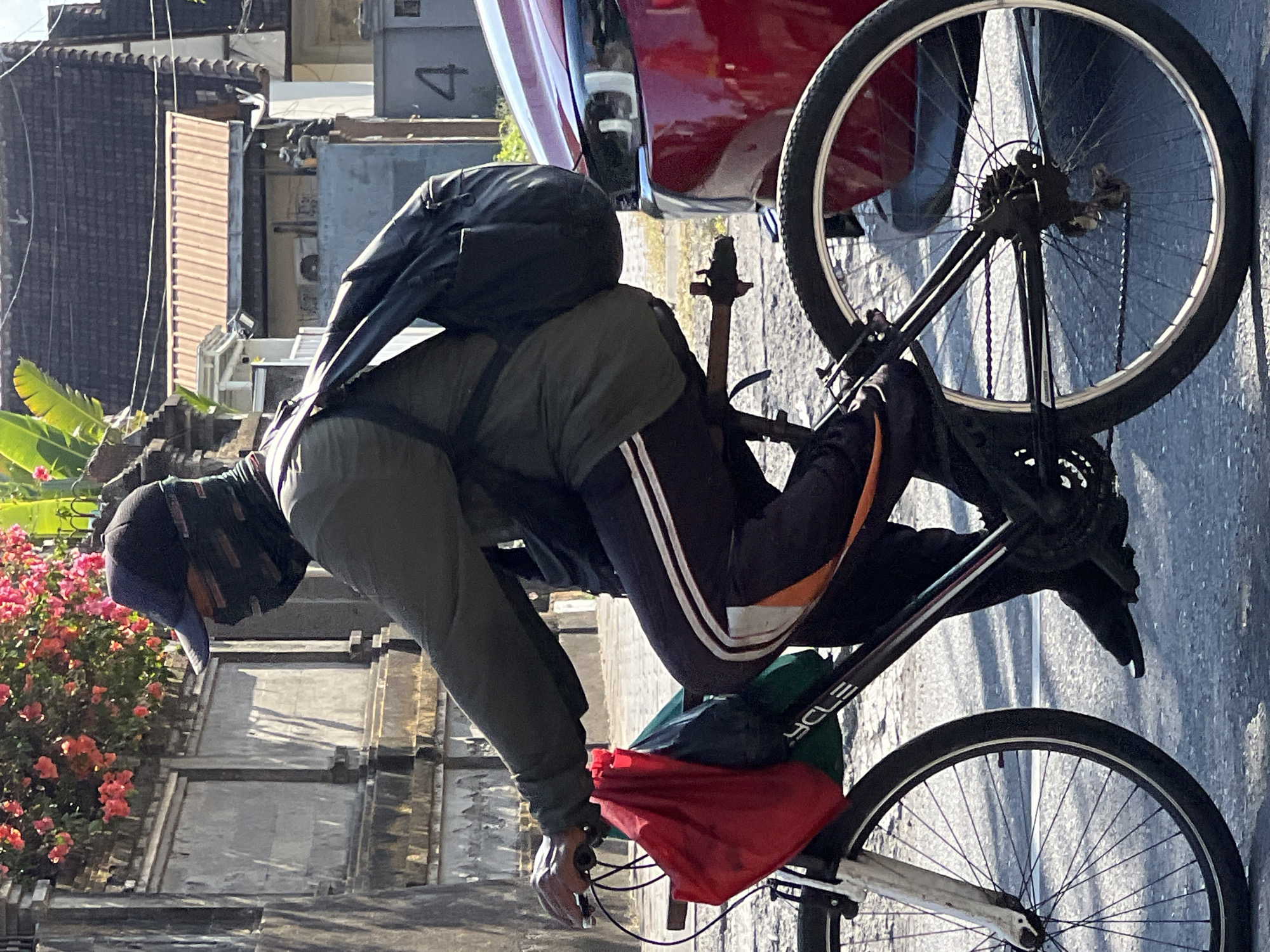 A person wearing a dark baseball cap and a black and grey face covering is riding a bicycle on a paved road in Denpasar, Indonesia. The cyclist is wearing a dark green long-sleeved shirt and dark pants with white and orange stripes down the sides. They have a black backpack on their back and a red bag and a green bag hanging from the handlebars of the bicycle. The bicycle is black with white rims on the wheels. In the background, there are buildings with tiled roofs, a red car, and stone walls with flowering plants. The scene appears to be in an urban or semi-urban environment.