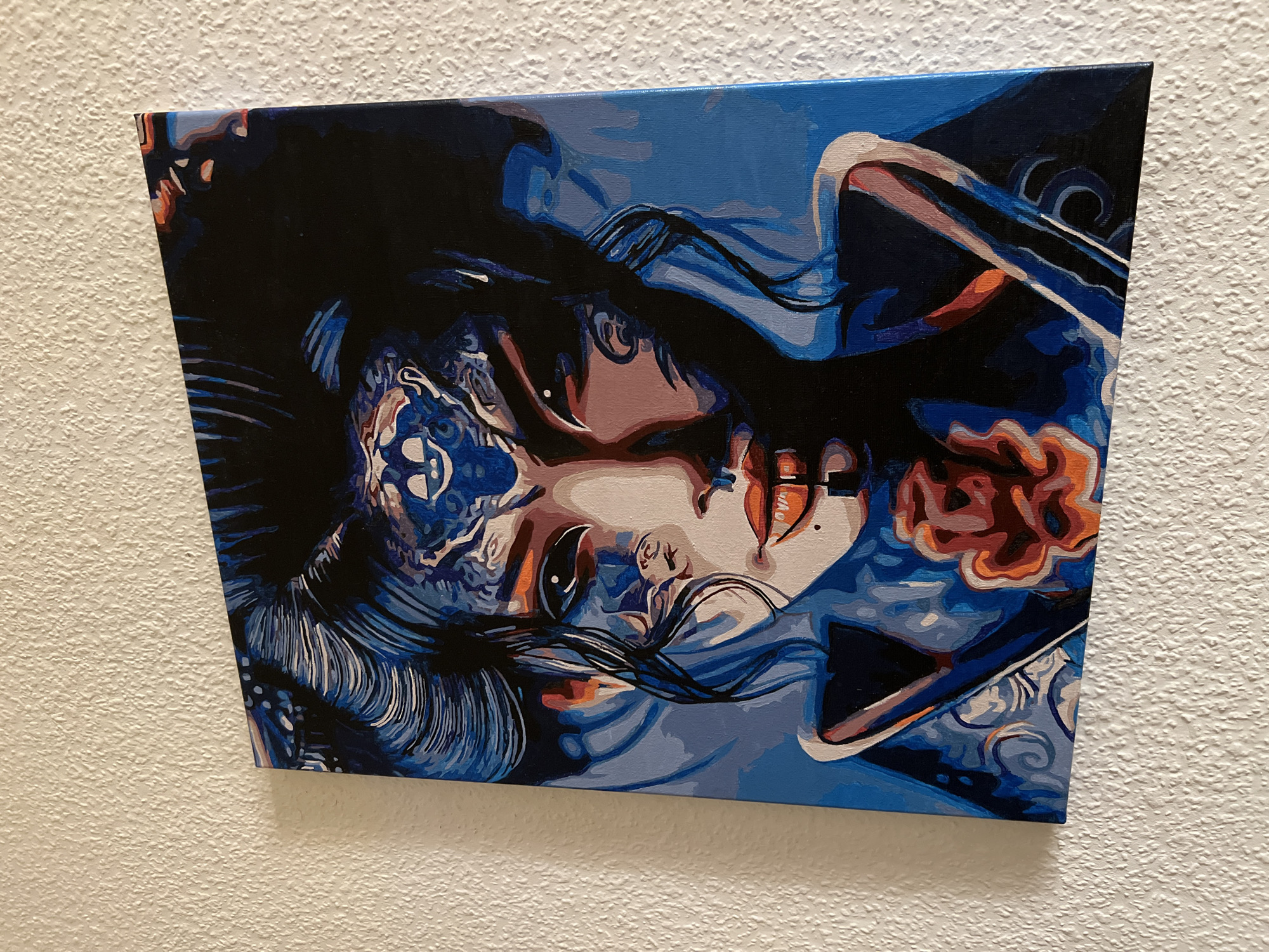 The image displays a colorful painting of a woman with intricate blue and orange designs on her face. The painting is mounted on a textured white wall. The woman has dark hair styled in an updo and her eyes are a striking black. Her lips are painted a bright orange, and there is a subtle dark line dividing them. A large, stylized orange and red flower is depicted on her chest, just below her chin. The background of the painting is a solid blue. The painting is a portrait, rendered in a stylized, almost graphic, manner.