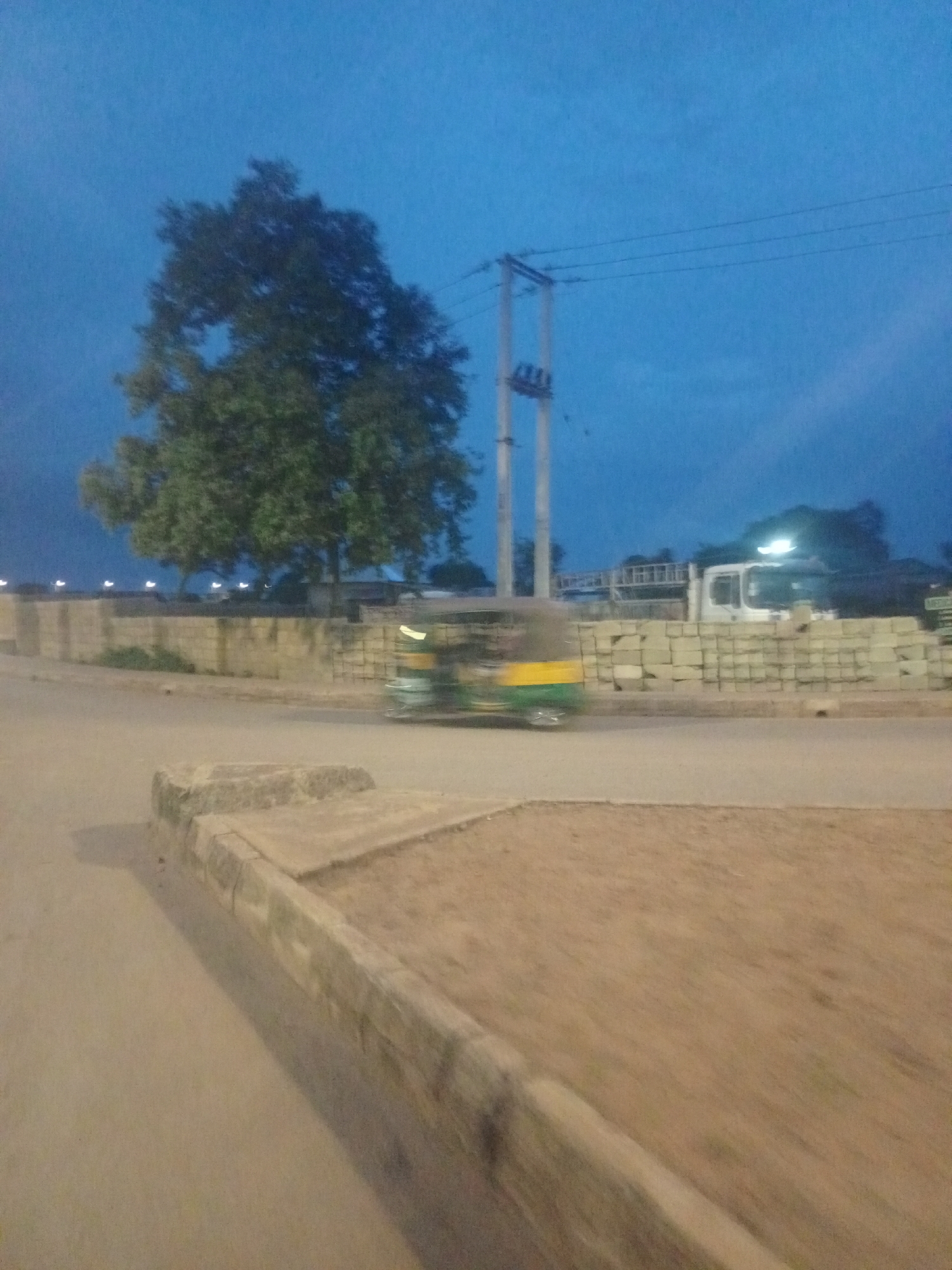 The image captures an outdoor scene at dusk in Bukuru, Nigeria. The sky is a deep blue, indicating the transition from day to night. In the foreground, a sandy, uneven surface with a raised concrete curb dominates the lower portion of the frame. To the left of the curb, a paved road is visible, blurred by motion, suggesting the photograph was taken from a moving vehicle.
Further into the scene, a brick wall runs horizontally, serving as a divider. Behind the wall, a large tree with dense green foliage stands prominently. To the right of the tree, a white truck with a ladder rack is parked, its headlights illuminated. In front of the truck and wall, a three-wheeled auto-rickshaw is in motion, blurred due to its speed. The auto-rickshaw has a yellow and green canopy. A series of concrete blocks are stacked neatly behind the brick wall, near the truck. In the background, faint lights are visible, possibly streetlights or lights from buildings. Electrical poles and wires traverse the upper portion of the sky, with one pole appearing to be a utility pole with transformers. The overall impression is of a roadside scene in an urban or semi-urban area during twilight.