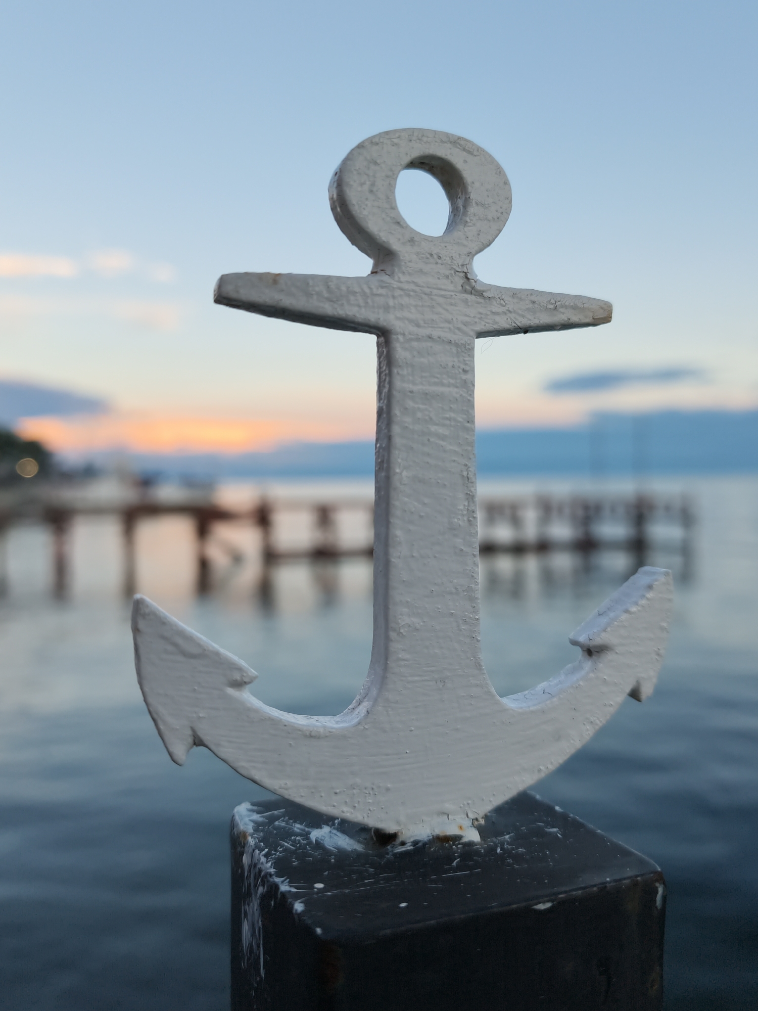 A close-up, vertical, eye-level shot features a white anchor ornament in the foreground, mounted on a dark metal post. The anchor is slightly worn, with peeling paint. The background is blurred, depicting a body of water, possibly a lake, with a pier or boardwalk extending into it. The sky above shows a gradient of pale blue with hints of pink and orange from a sunset. The location is specified as Listvyanka, Russia. There are no people or animals visible in the image.