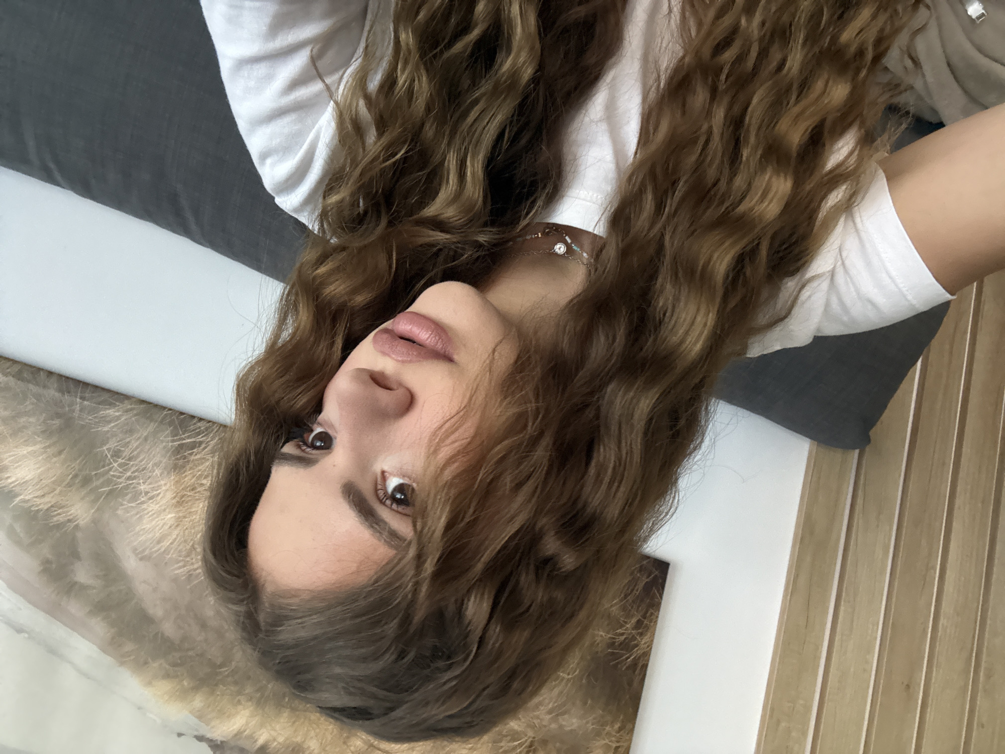 A woman with long, wavy brown hair and light skin is seen in a close-up, angled shot. She wears a white top and a delicate necklace. Her eyes are brown, and her lips are a neutral color. The background includes a gray fabric, possibly upholstery, and a wall with vertical wooden panels. The location is Unknown, Unknown.