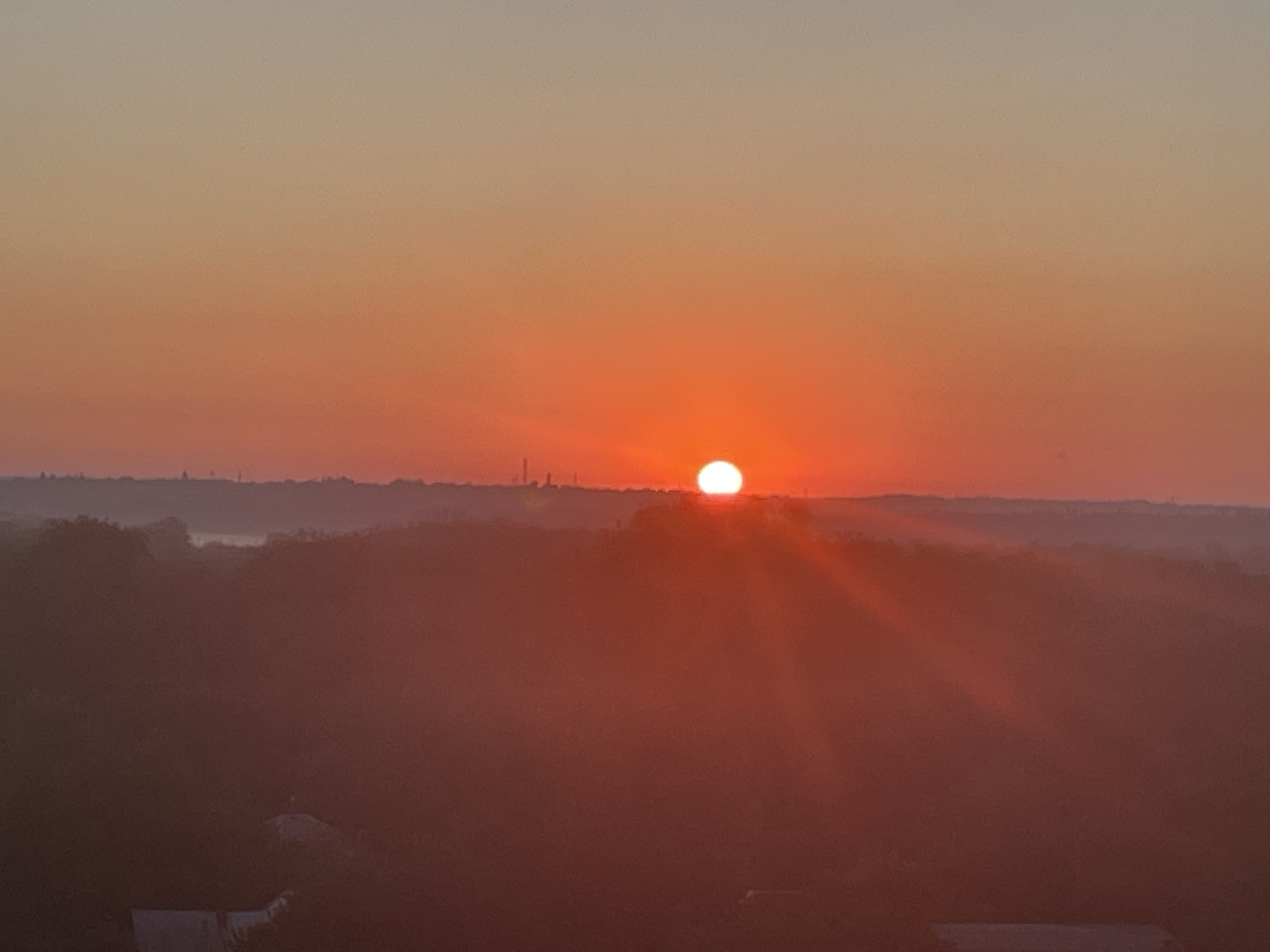 This image depicts a sunset over Novocherkassk, Russia. The sun is a bright, orange orb near the horizon, casting a warm glow across the sky which transitions from a pale yellow at the top to a deep orange closer to the horizon. The landscape below is obscured by a reddish haze, suggesting either mist or dust. Silhouetted against the vibrant sky are rolling hills and distant structures that appear to be buildings and possibly industrial elements. Some trees are visible as dark shapes on the hillsides. In the foreground, the haze is thicker, making details indistinct, but some faint shapes that could be rooftops of houses are discernible at the bottom of the frame. The overall impression is one of a serene and atmospheric end to the day.