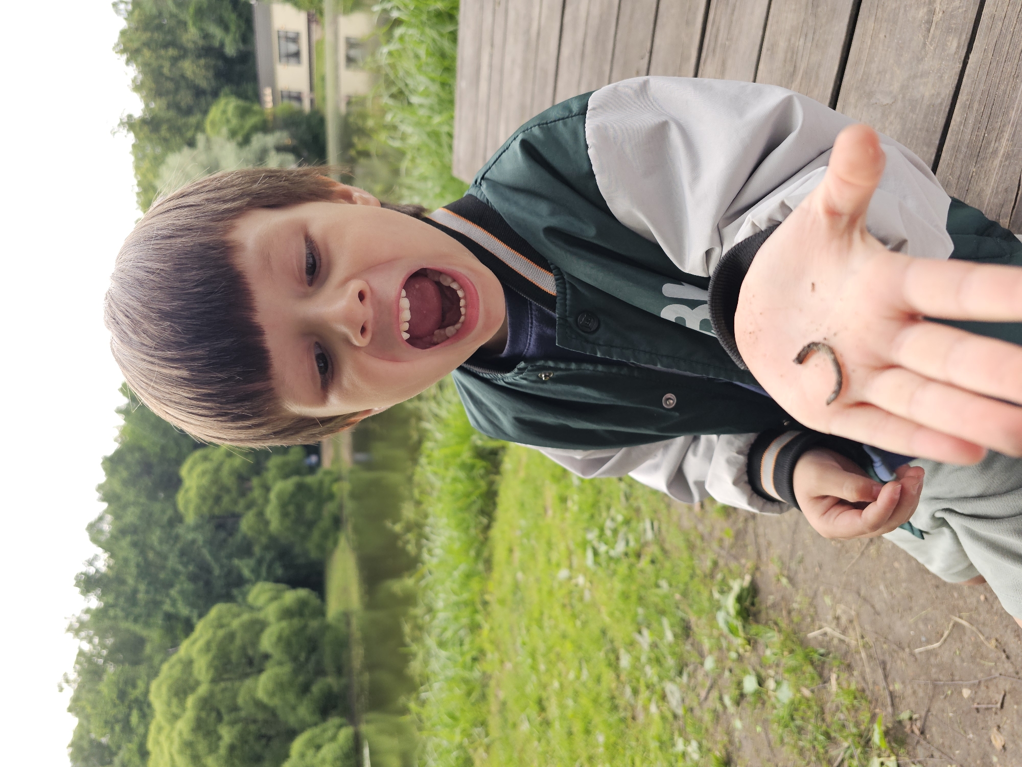 A young boy, with brown hair and an open mouth, is sitting on a wooden boardwalk outdoors. He is holding a small, dark worm in his open palm, displaying it to the viewer. The boy is wearing a dark green and grey jacket with white sleeves and orange and black trim around the collar and cuffs. Behind him, a body of water reflects lush green trees and a building. The scene appears to be a park or natural area, possibly within the city of Moscow, Russia, given the presence of trees, water, and a building in a park-like setting.