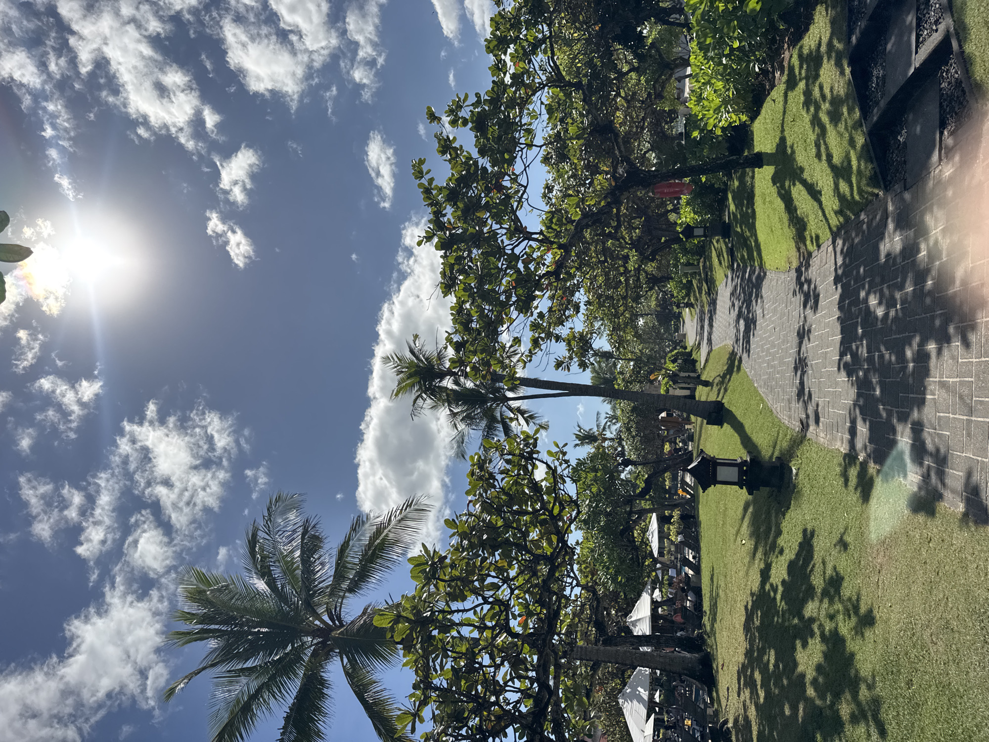 The image depicts a tropical landscape under a bright, sunny sky in Kuta Selatan, Indonesia. The sky is blue with scattered white clouds and a visible sun glare in the upper left corner. The scene unfolds along a paved walkway lined with trees, including prominent palm trees. Lush green grass flanks the walkway. The shadows cast by the trees are visible on the grass and walkway, indicating strong sunlight. In the distance, possibly an outdoor seating area, there are white umbrellas providing shade. The photograph suggests a tranquil, possibly resort-like or park-like setting. There are no visible people in the main scene, though there might be obscured figures near the distant umbrellas.
