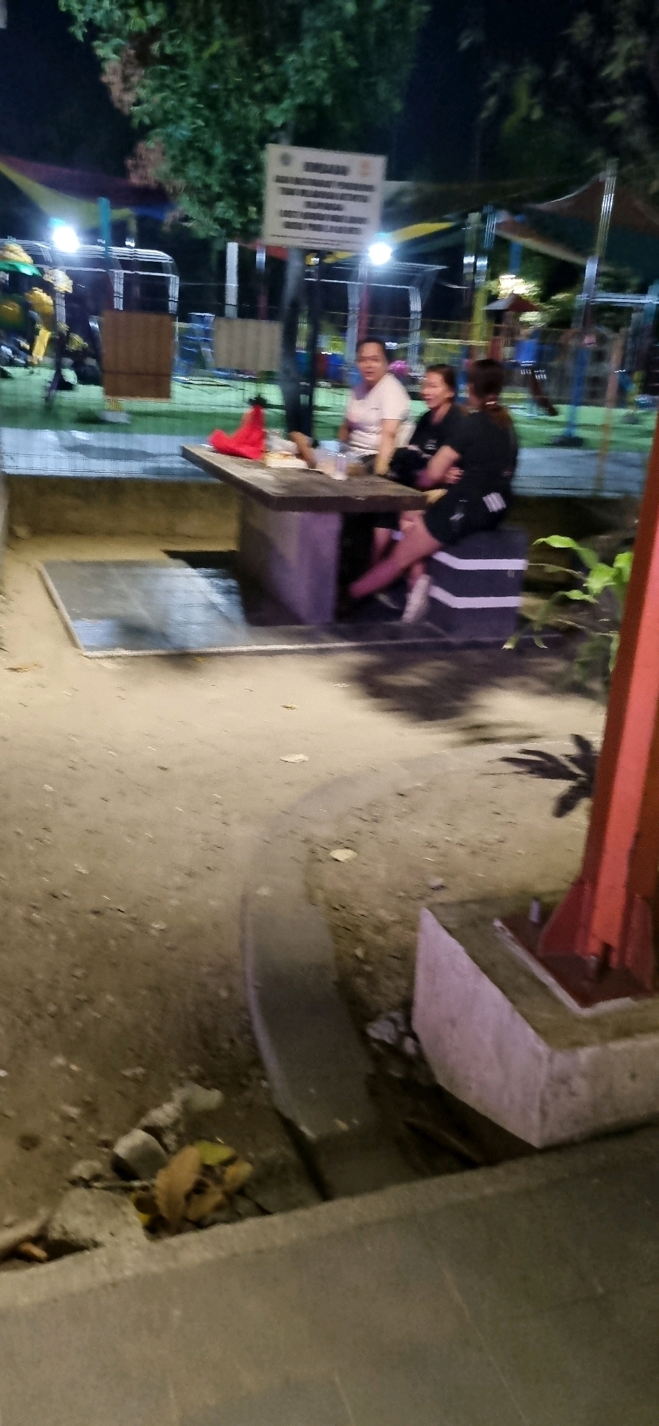 The image shows a group of people sitting around a concrete picnic table at night. The setting appears to be an outdoor recreational area, possibly a park or a public space. In the background, there are some colorful awnings and playground equipment, suggesting it might be a children's play area. There is also a sign post visible, but the text on the sign is illegible. The ground is covered in dirt and gravel. The lighting is dim, indicating it is nighttime, and there are some artificial lights illuminating the area. The people are casually dressed, and their interactions are not clear from the image. The presence of picnic tables and playground equipment suggests this location is in a public area where people gather for recreation. While the exact location in Denpasar, Indonesia, is not specified in the image, such an environment is common in urban parks or community recreation zones within the city.