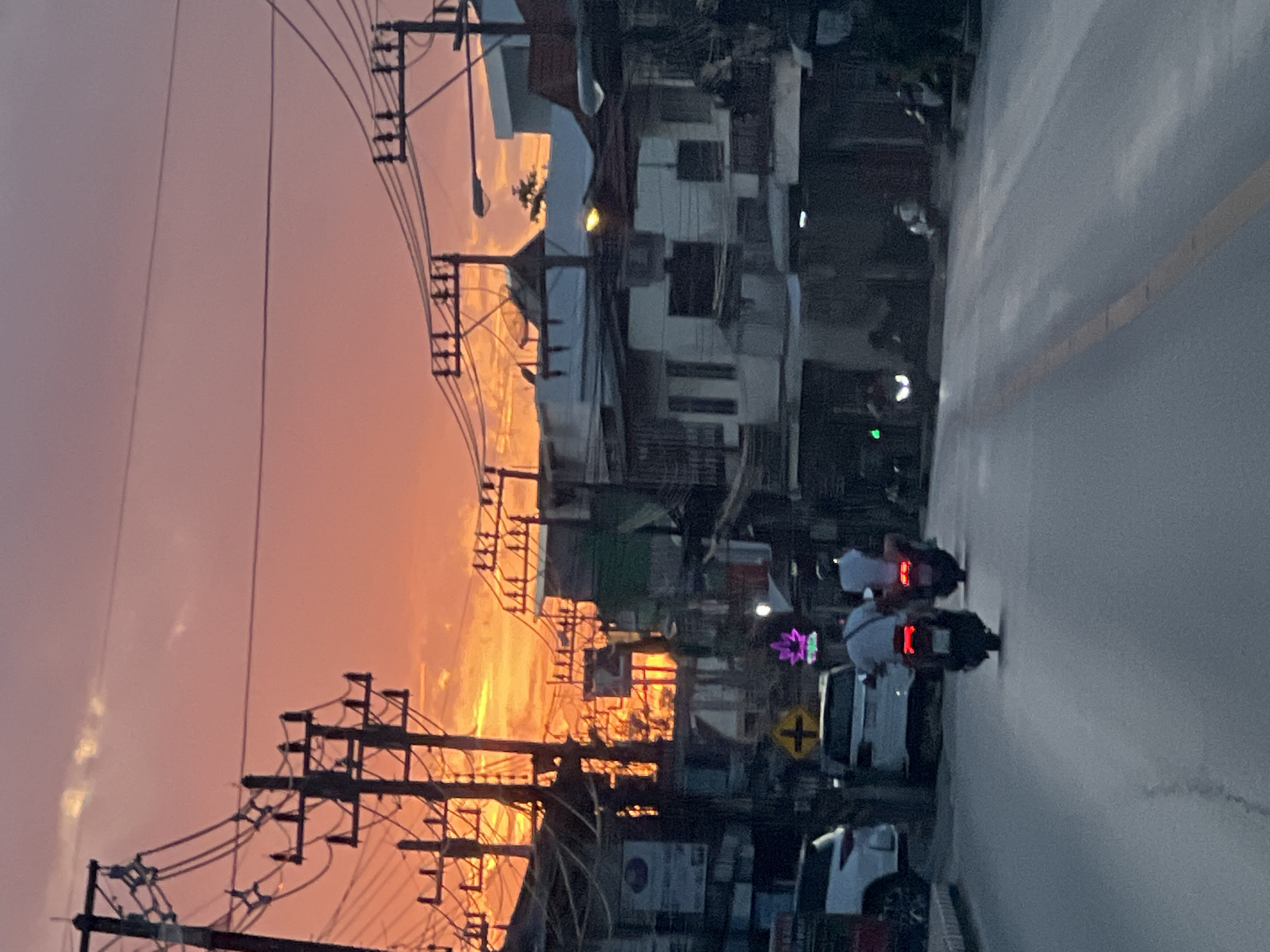 The image depicts a street scene at dusk in Baan Mae Nam, Thailand. The sky is filled with the vibrant colors of a sunset, ranging from deep orange to pink and purple hues. Electrical poles with numerous wires crisscross the upper portion of the frame, silhouetted against the colorful sky. In the foreground, a paved road with a yellow line marking the center runs towards the background. Several vehicles are visible, including two white cars and two motorcycles carrying riders. The motorcycles are in motion, with their brake lights illuminated. Buildings line the street, appearing to be a mix of residential and commercial structures, with some showing signs of wear and tear. The overall atmosphere is that of a typical Southeast Asian street scene at the end of the day, with the warm glow of the sunset illuminating the surroundings.