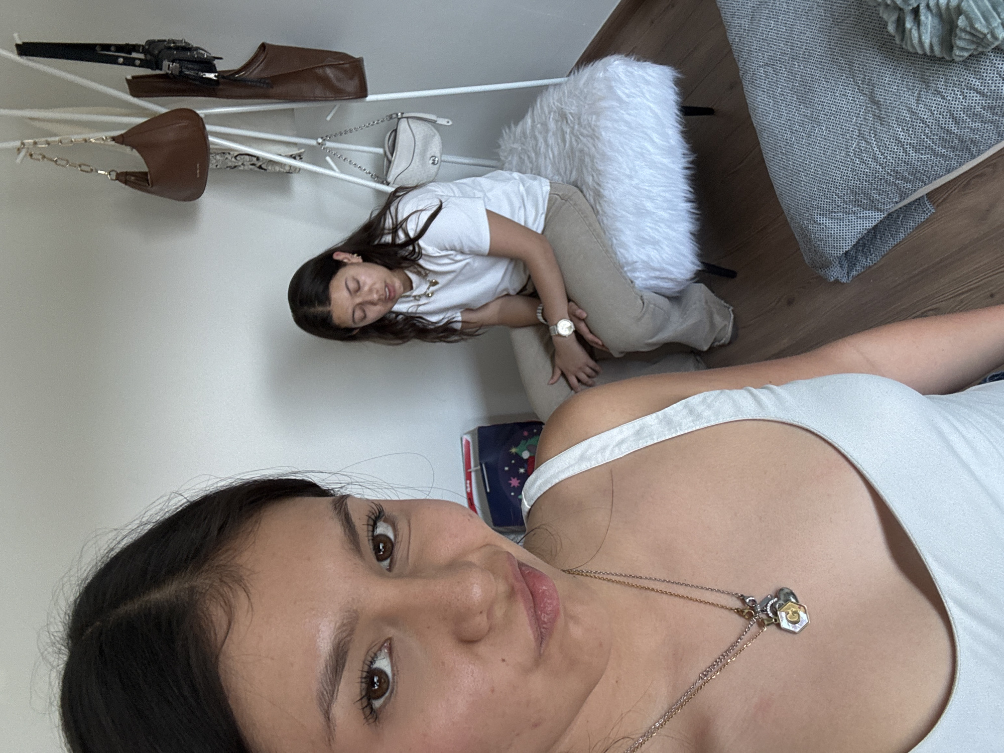 The image, taken in an indoor setting, features two young women and various personal items. The photo is rotated 90 degrees clockwise, making the subjects appear on their sides.
In the foreground, a young woman with dark hair and brown eyes looks directly towards the camera, suggesting a selfie or direct gaze. She wears a white tank top and multiple necklaces, including one with a gold-colored hexagonal charm and another with a small heart pendant. Her expression is neutral with slightly parted lips.
In the background, another young woman with dark hair is seen leaning or lying on a white, fluffy stool or chair. She is wearing a white t-shirt, light-colored pants, and a watch on her left wrist. Her eyes appear closed or downcast, giving a relaxed impression.
Above the background subject, a white rack or shelf holds several items: a brown, soft-structured purse with a chain strap, another off-white/silverish purse, a dark object resembling a weapon or toy gun, and a brown shoe or slipper. The room features a light-colored wall and a wooden floor. Part of a bed or seating area is visible in the upper right, covered with a gray patterned blanket and a greenish textured throw. A blue box with a festive design is partially visible near the background subject's legs. No distinct text is visible on any objects or surfaces. The overall atmosphere is casual and personal, likely within a bedroom or living space.