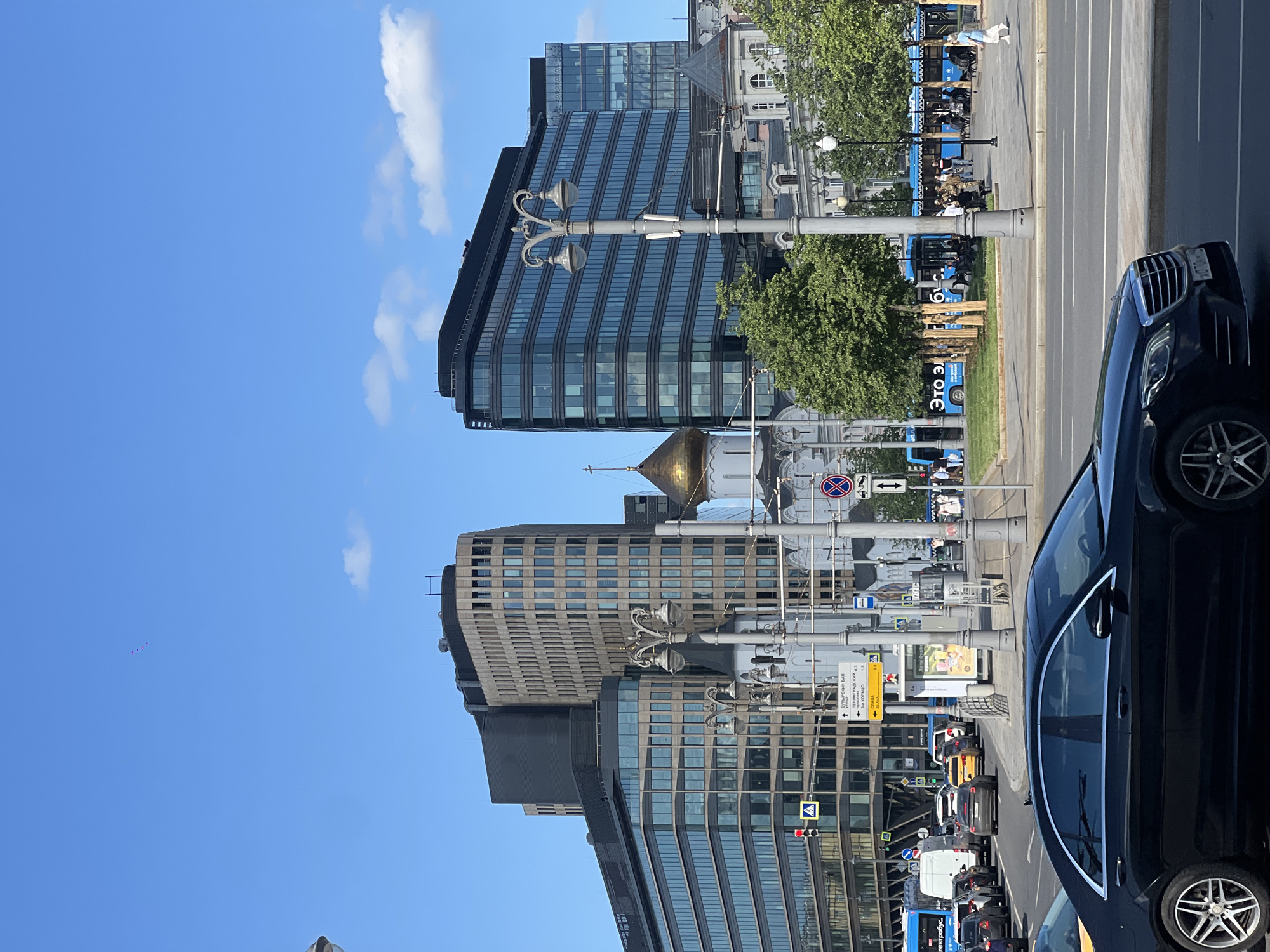 The image captures a street scene in Moscow, Russia. In the foreground, a black Mercedes-Benz passenger car is parked. Further into the frame, a line of cars, including a white van, follows the road. The scene is marked by its architecture, featuring several modern office buildings with glass facades. A golden-domed building, most likely a cathedral or church, is visible behind the office buildings. Street lights are aligned along the road. On the sidewalk, people are seen walking and sitting on blue public benches. The sky is mostly clear, with a few clouds. There are signs indicating traffic and directions to various locations, including Leningradskii Prospekt. The overall atmosphere is urban, representing the city life.