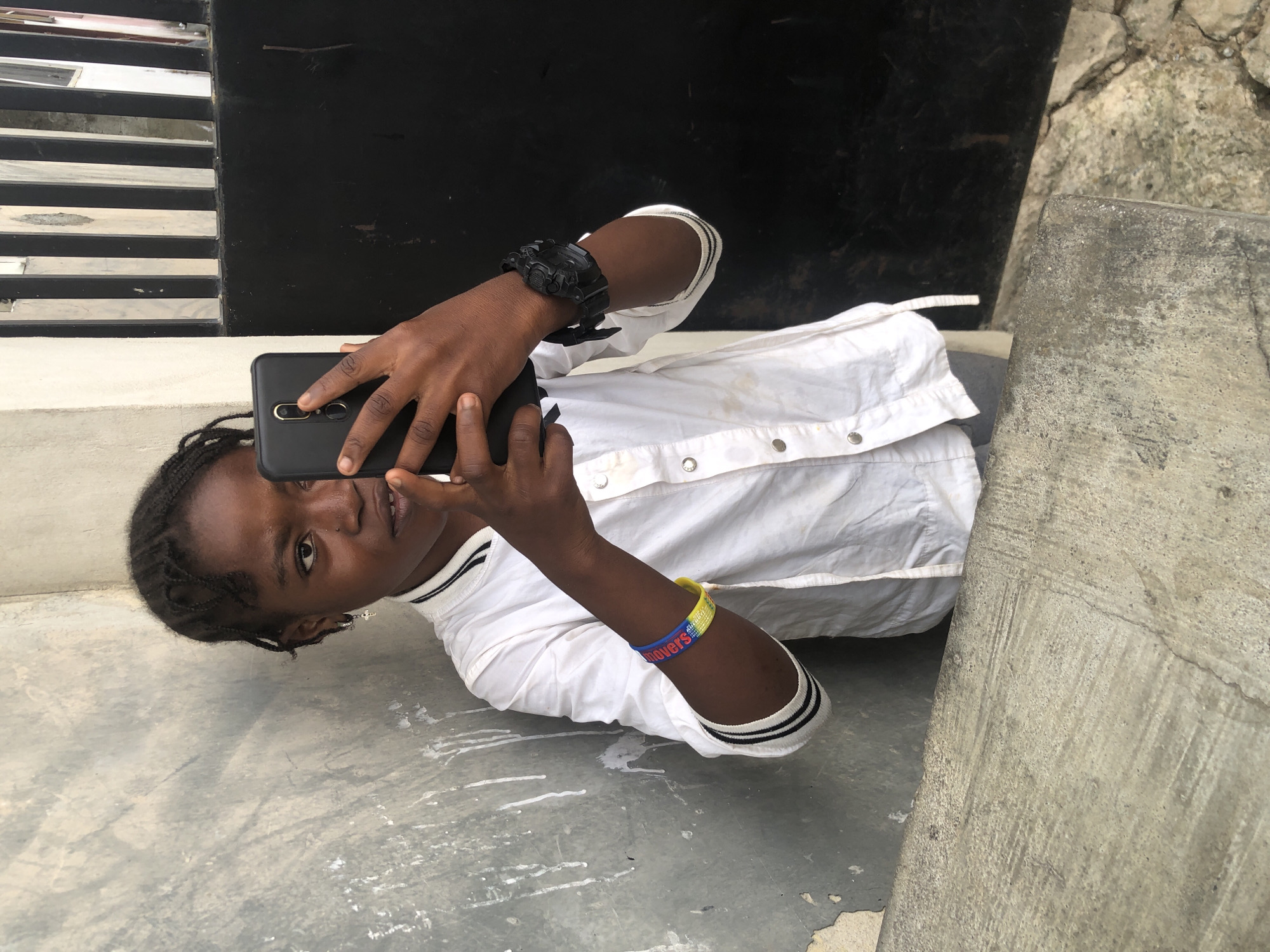 A young Black girl with braided hair and a white collared shirt with black stripes on the cuffs is standing outdoors in Ado, Nigeria, and taking a selfie with a black smartphone. She is wearing a silver cross earring in her left ear, a colorful bracelet on her left wrist, and a black watch on her right wrist. The background consists of a rough concrete wall on the left and a black gate with vertical metal bars on the right. A concrete ledge is in the foreground. The image appears to be taken during the daytime.