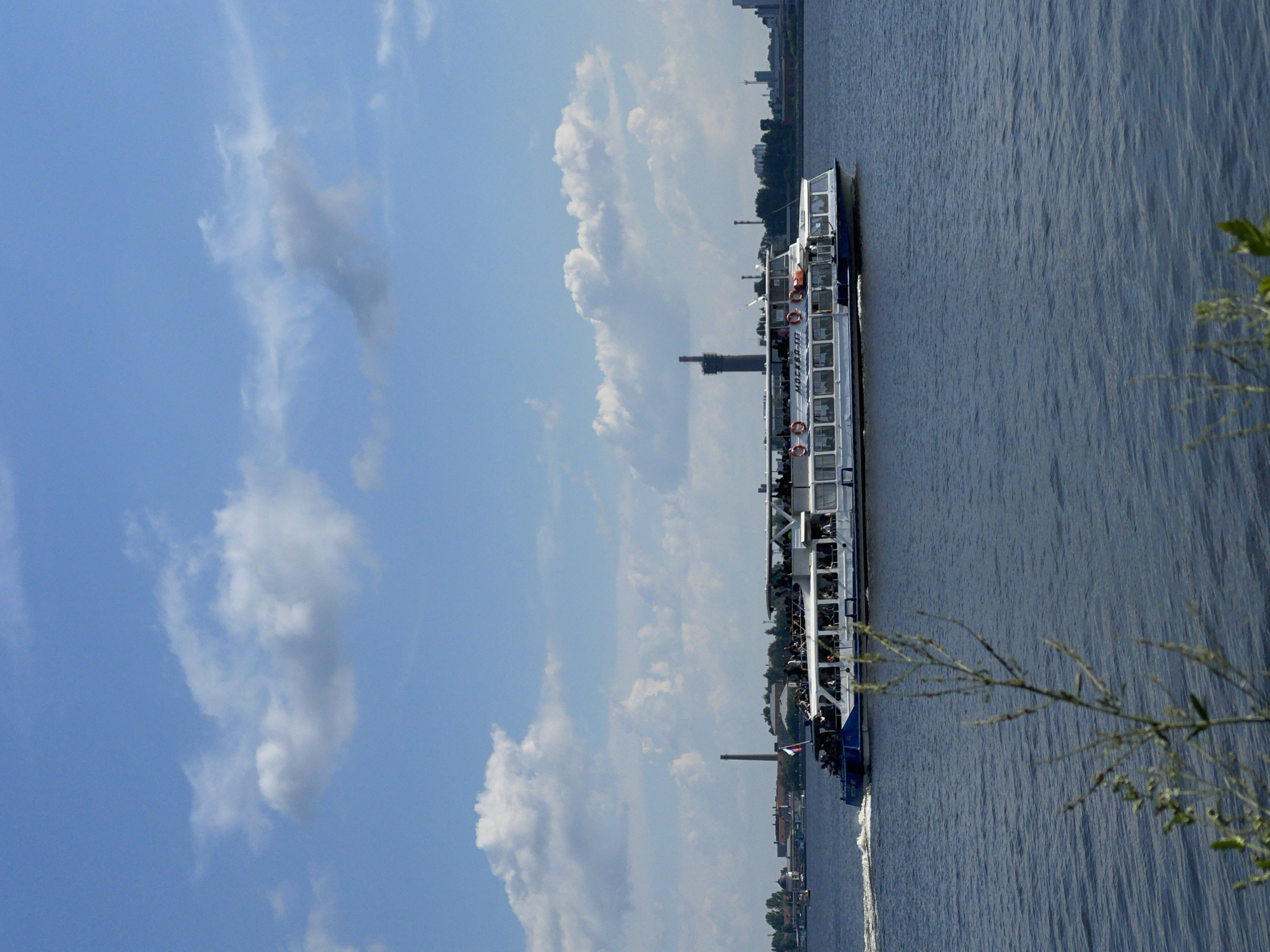 The image shows a multi-deck passenger boat navigating a body of water, likely a river, with a city skyline in the background under a blue sky with scattered clouds. The boat, labeled "МОСКВА-117" in Cyrillic, is white with blue trim and appears to be carrying a significant number of passengers on its open upper deck and enclosed lower decks. Life preservers are visible on the side of the upper deck. The city in the background features industrial structures, including a tall smokestack, and various buildings, suggesting an urban or industrial area. Vegetation is present along the shoreline. The overall scene depicts a typical day of riverine transportation and urban activity. While the image's metadata is not available to confirm the exact location, the inscription on the boat "МОСКВА" (Moskva) suggests it may be a vessel operating in or related to Moscow. However, it is not possible to definitively place this image in Izhevsk, Russia, based solely on the visual information provided.