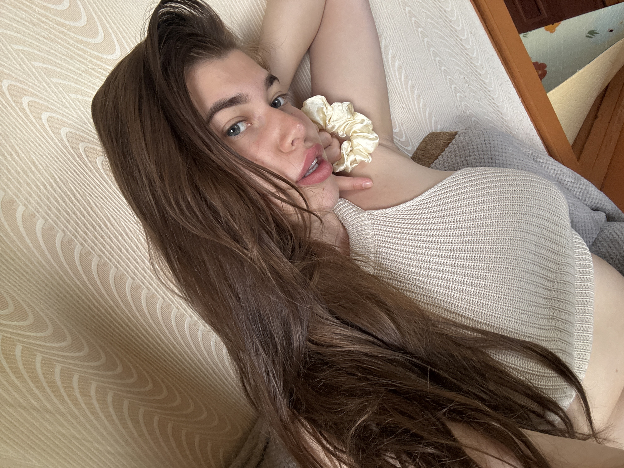 The image shows a young woman with long, dark brown hair and blue eyes, lying on her side on a textured, beige surface. She is wearing a light-colored, ribbed tank top. Her right arm is bent, and her hand is resting on her cheek. A light yellow scrunchie is visible on her forearm. The background is a beige wall with a subtle, wavy pattern. The scene appears to be indoors, possibly in a bedroom, given the soft lighting and relaxed pose. There are no identifiable landmarks or features that would suggest a specific location within the city of Мещёры, Russia. The focus is on the woman and her immediate surroundings.