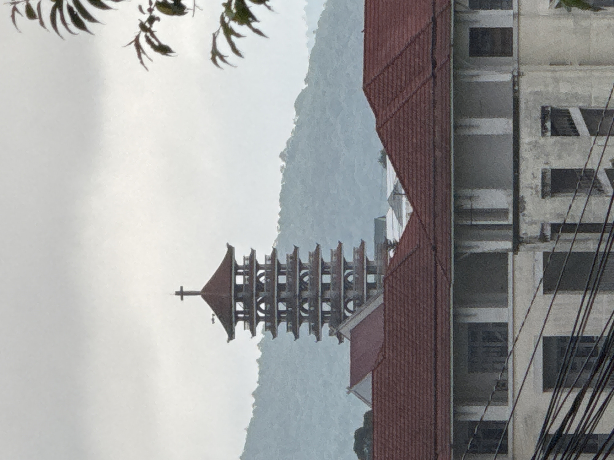 This image captures a serene, hazy view dominated by architectural structures set against a mountainous backdrop. The primary subject is a distinctive multi-tiered tower, likely a bell tower or a pagoda, crowned with a cross at its apex. This tower, with its numerous open levels and arched designs, emerges from a building featuring a red-tiled roof, visible towards the bottom of the tower.
To the left, a range of mountains is visible, appearing soft and indistinct due to atmospheric haze, mist, or fog, which gives the scene a diffused, quiet quality. The sky above is similarly overcast or hazy, contributing to the muted lighting.
In the right foreground, part of a multi-story concrete building is prominent. This structure exhibits several rectangular window openings, some appearing dark, and its surface shows signs of age or incomplete construction. A cluster of dark utility cables or wires diagonally spans across the facade of this building, adding a touch of urban reality to the scene. Another section of a red-tiled roof is also visible in the bottom right corner, suggesting more buildings out of frame.
Overhanging dark green foliage from a tree is visible in the top left corner, partially framing the upper left portion of the image. The overall impression is one of peacefulness, possibly a cool and humid day in a mountainous region. No people are visible, and there is no discernible text. Given the context, the architecture and misty mountains are characteristic of Da Lat, Vietnam, strongly suggesting the Da Lat Cathedral (Con Ga Church) as the central tower.