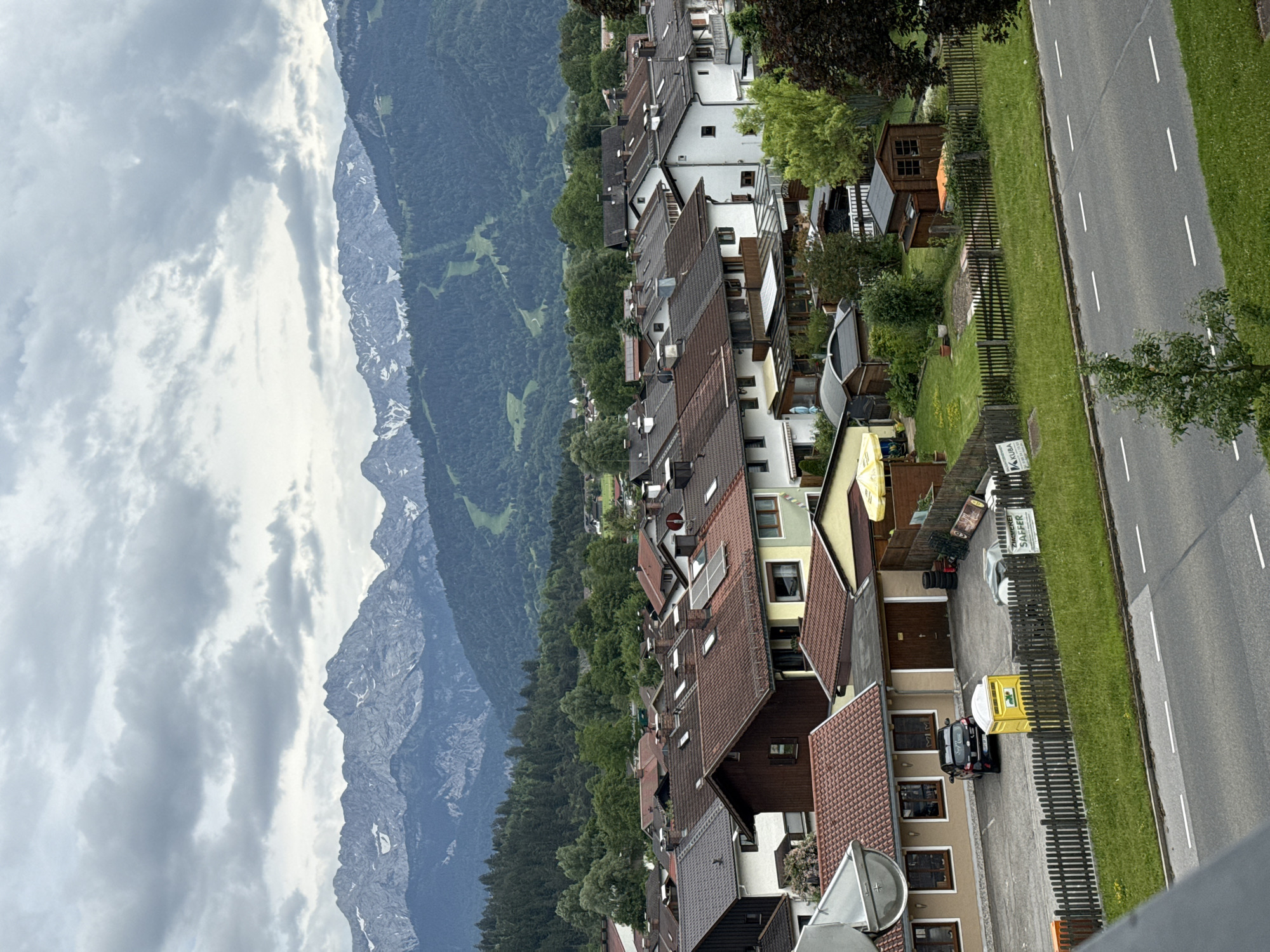 The image shows a suburban residential area in Garmisch-Partenkirchen, Germany, with a backdrop of the Bavarian Alps. In the foreground, a paved road with white lane markings runs across the bottom of the frame. Adjacent to the road is a strip of green grass and a wooden fence. Behind the fence are several residential houses with tiled roofs, some featuring solar panels. A black car is parked in front of one of the houses, next to a yellow portable toilet. Further back, there are more houses and trees, with the foothills of the Alps rising behind them. The mountains themselves are prominent in the background, their peaks partially covered in snow, and the sky is overcast with gray clouds.