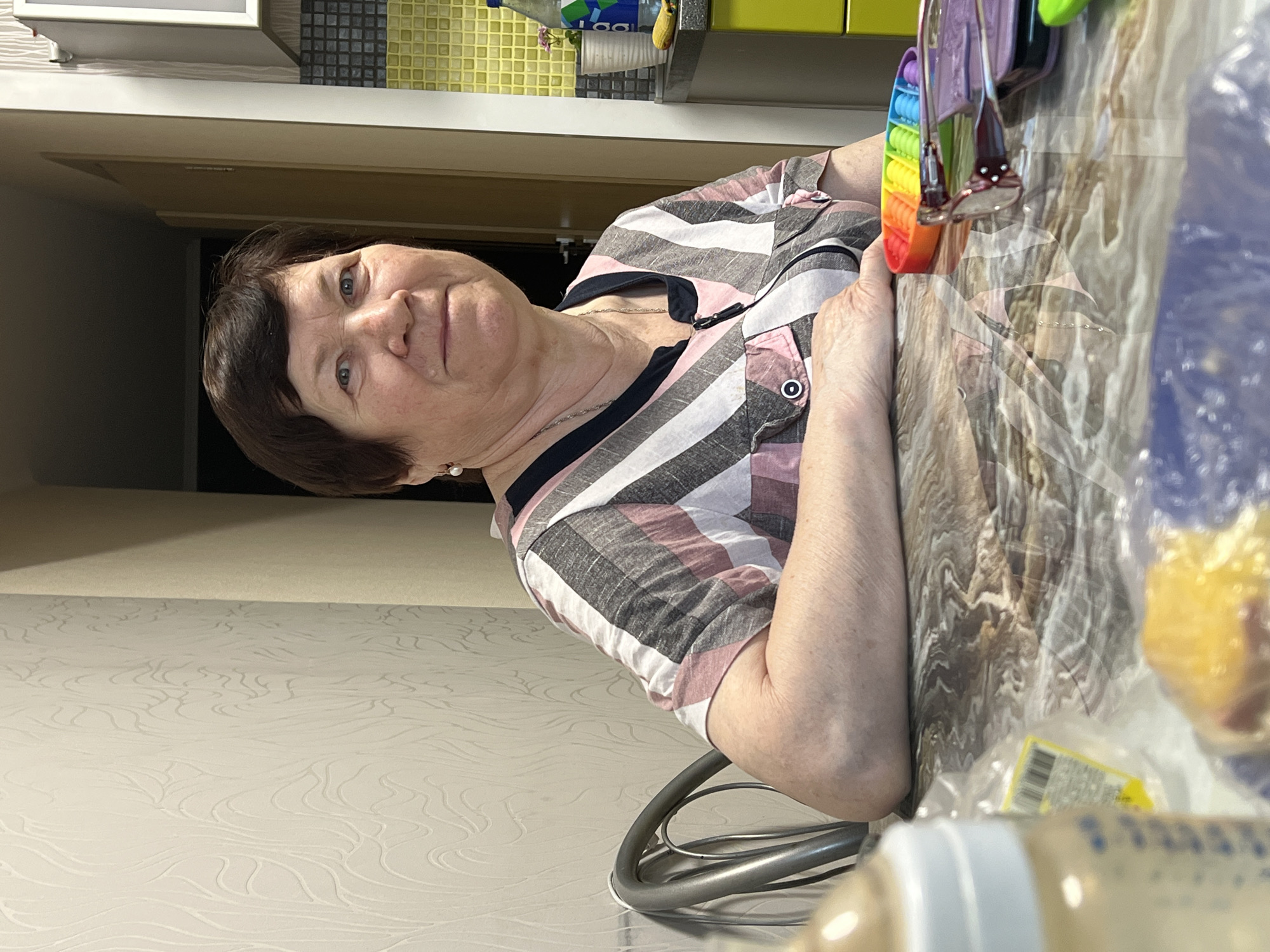 The image features an older woman seated at a table in what appears to be an indoor setting, likely a home kitchen or dining area. She has short, dark brown hair and blue eyes, looking directly at the camera with a neutral to slightly pleasant expression. She is wearing a short-sleeved, collared shirt with distinct stripes in white, light pink/mauve, and dark grey/black. A thin silver chain necklace and a small pearl earring in her left ear accessorize her outfit. Her forearms rest on the table surface.
On the table in front of her right arm is a vibrant rainbow-colored "Pop It" fidget toy. The table surface itself appears to be covered, revealing a light-colored, possibly wood-grain or abstract textured pattern underneath. In the background, a light-colored wall features a subtle, swirling patterned wallpaper. Above this, a tiled backsplash combines yellow and black/grey square tiles. A shelf or countertop above the backsplash holds a partially visible bottle of drink with a blue and white label, where some text resembling "LAAL" or similar letters can be discerned. A small decorative plant or object is also on the shelf.
In the blurred foreground on the right, several items are visible, including a plastic bag with yellow contents, a baby bottle with a white cap and blue markings containing a light brown liquid, and a blurry yellow label with a barcode. The woman is seated on a chair with a distinctive curved metal frame. The lighting suggests an indoor environment, creating a candid portrait.
