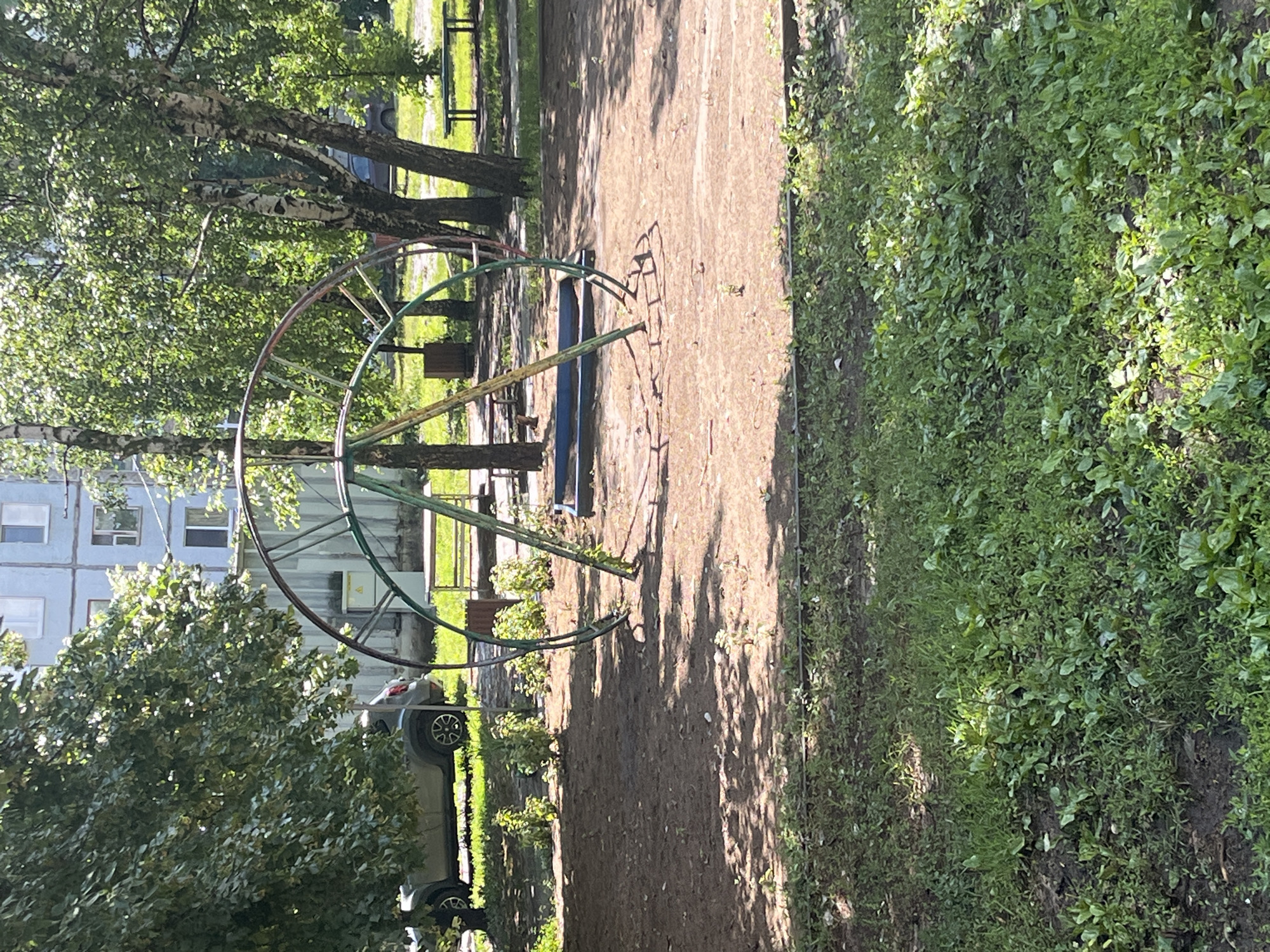 A playground merry-go-round with a metal frame in green, red, and yellow is located on a dirt surface. A blue plastic seat is visible beneath the structure. In the background, a light blue apartment building and several trees with green leaves are present. The ground is covered with dirt and some patches of grass. A silver car is parked to the left of the merry-go-round, and two wooden benches are visible in the distance. The scene is illuminated by sunlight, creating shadows on the ground.