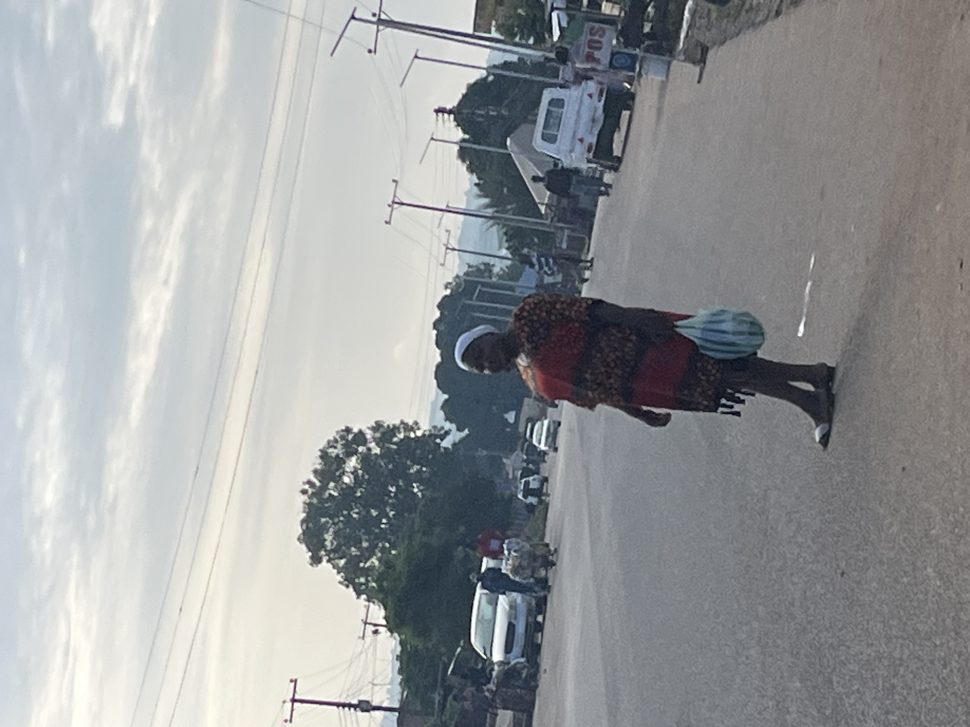 A woman in a red and black patterned dress and a white head covering walks across a paved road. She is carrying a striped blue and white bag in her right hand. Her feet are bare except for what appear to be flip-flops or sandals. The road appears to be a commercial area in Jos, Nigeria, with utility poles and street lights lining the sides of the road. Several vehicles are present, including a white car, a white pickup truck, and other cars in the distance. Other people are visible on the sides of the road, and some are pulling carts. The sky is overcast with clouds.