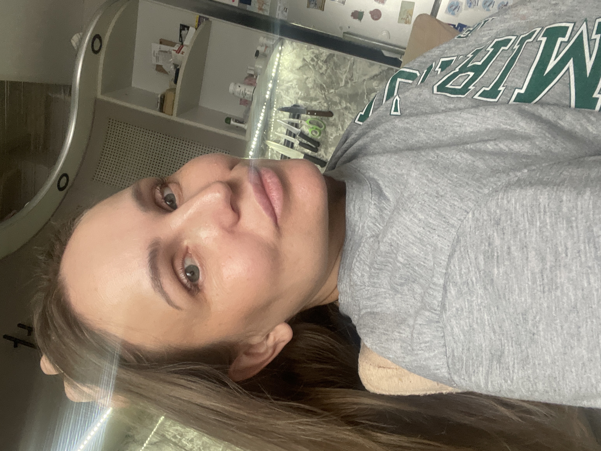 A young woman is captured in a close-up, portrait-style shot indoors in Chaikovsky, Russia. She has light brown hair and is wearing a grey marl sweatshirt with a partial green and white text visible, likely spelling "CHAMPION." Her head is tilted, and she looks directly into the camera with a neutral expression.
The background reveals a domestic setting. To her left, part of a circular mirror or decorative object is visible, along with a white shelved cabinet containing various bottles and miscellaneous items. Below the cabinet, a white radiator or vent is present. To her right, a countertop or shelf is visible, featuring several kitchen knives with different handles (some wooden, some black, some white), and a pair of green-handled scissors. This area is illuminated by a strip of artificial light, possibly under-cabinet lighting, creating a bright reflection. The overall lighting suggests an indoor setting, potentially during the evening or night. The image gives a casual and candid feel of the woman in her personal space.