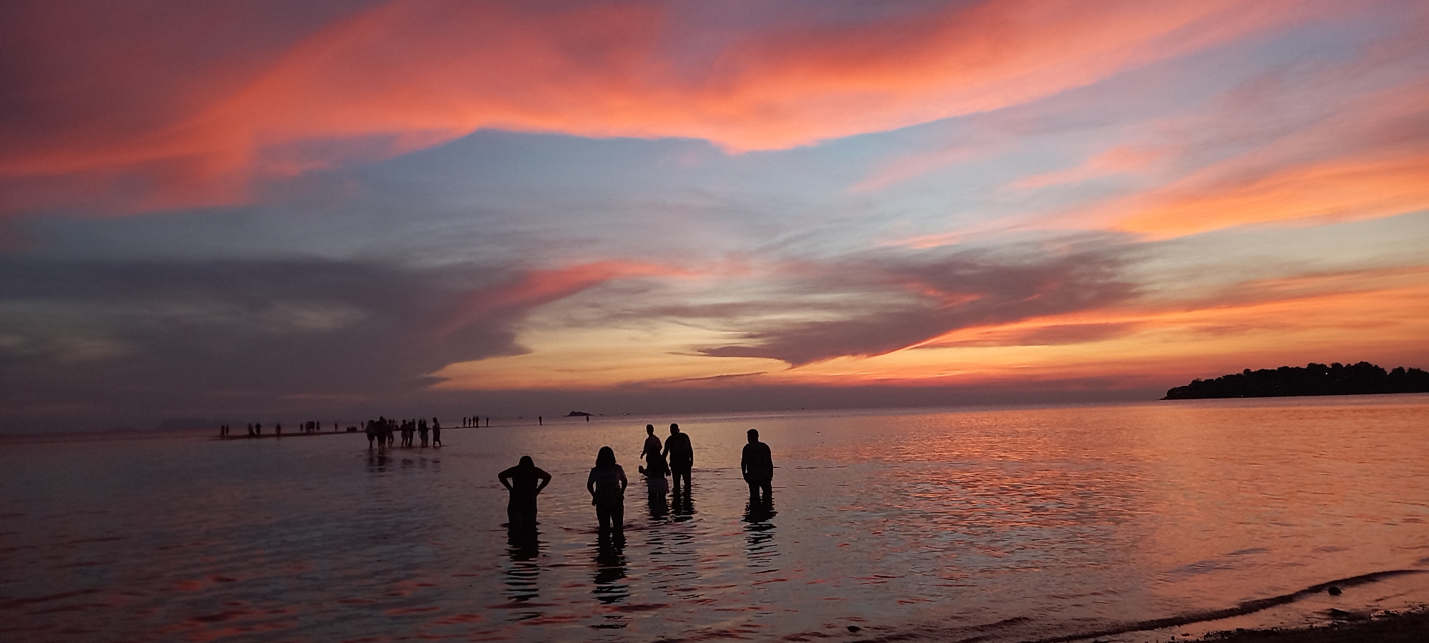 The photograph captures a sunset at Baan Sri Thanu, Thailand. Silhouetted figures are wading in shallow water, while others stand further away on a sandbar. The sky is painted with streaks of pink, orange, and gray clouds. A dark, tree-covered island is visible on the right horizon. Ripples are reflected across the water's surface.