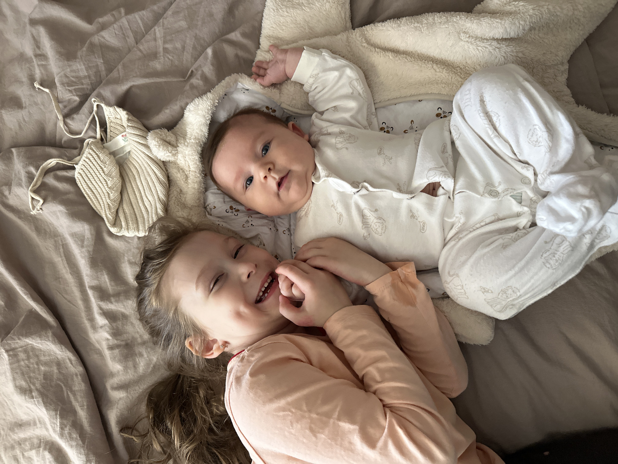 A high-angle shot shows two children, a baby and a young girl, lying on a bed. The baby, dressed in a white onesie, is lying on a white blanket with a light-colored, fluffy border. The girl, wearing a peach-colored long-sleeved shirt, is lying next to the baby with her hands clasped together near her face, smiling with her eyes closed. A light-colored knitted hat is lying on the bed to the left of the baby. The bed is covered in a light gray sheet. The location is Chaikovsky, Russia.