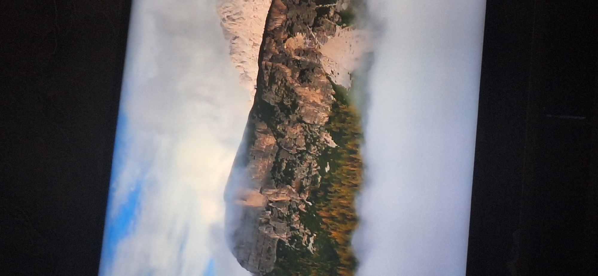 The image shows a mountain range partially obscured by clouds and fog. The mountains are rocky with some sparse vegetation, including trees with yellow and orange foliage, suggesting an autumn season. The sky above is partly cloudy with patches of blue. The foreground is filled with a thick layer of white fog or clouds. There is no indication of the city of Bila Tserkva, Ukraine, in this image.