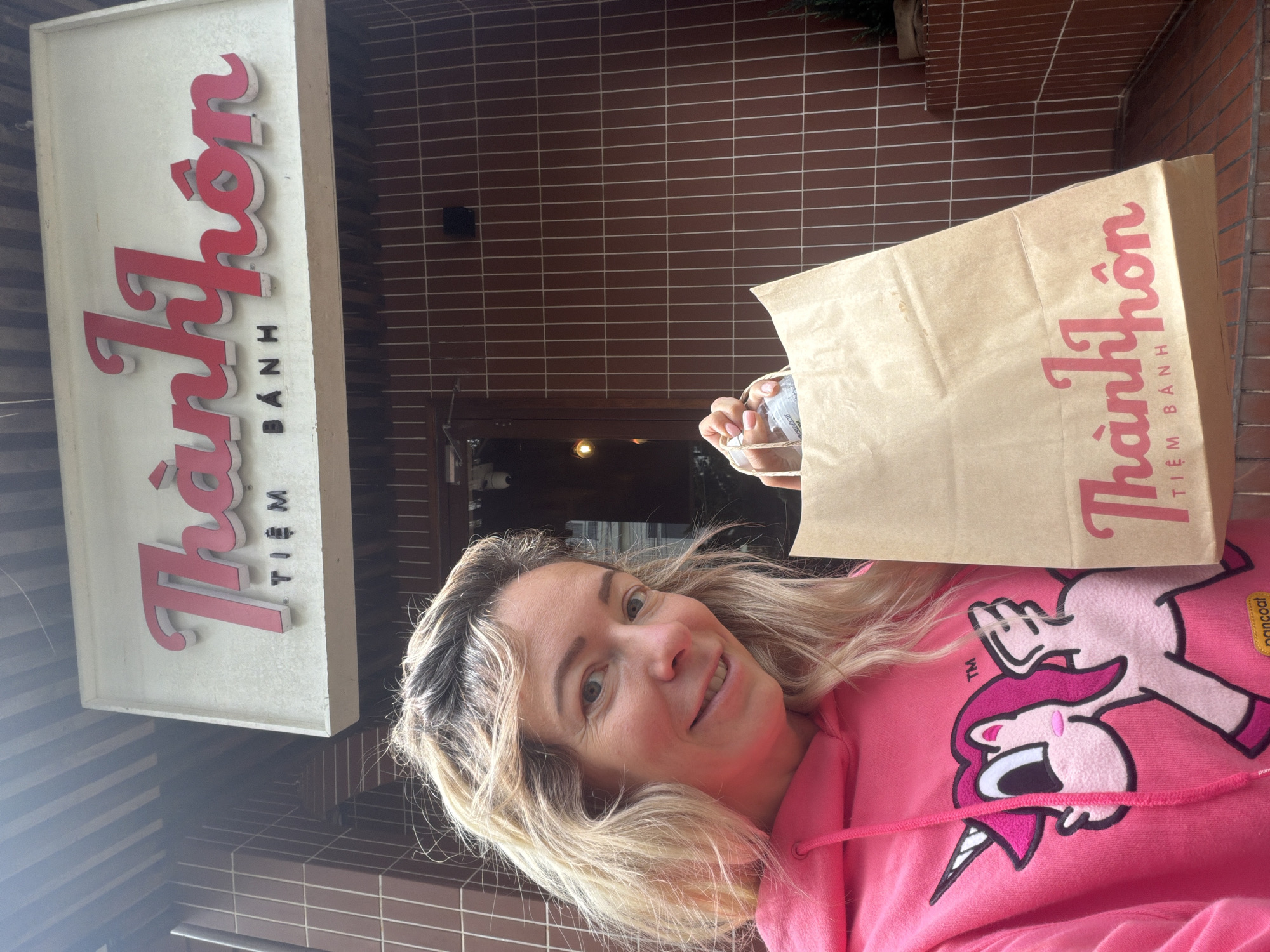 A woman with blonde hair, wearing a pink hooded sweatshirt with a unicorn graphic, stands outside a building in Da Lat, Vietnam, holding a brown paper bag. The building has a sign with red lettering that says "Thành Hôn Tiệm Bánh" and is clad in brown tiles. The woman is smiling at the camera. The environment appears to be an outdoor, urban setting, possibly a street or walkway, with an overcast sky.
