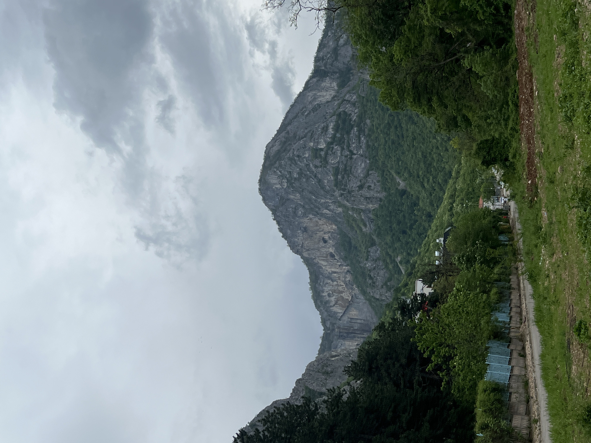The image shows a mountain landscape with a path running along the right side of the frame. The sky is overcast with grey clouds. The mountain is rocky and covered in some vegetation. There are houses and trees visible near the base of the mountain. The image is taken during daytime. The location is not specified in the image or the prompt.