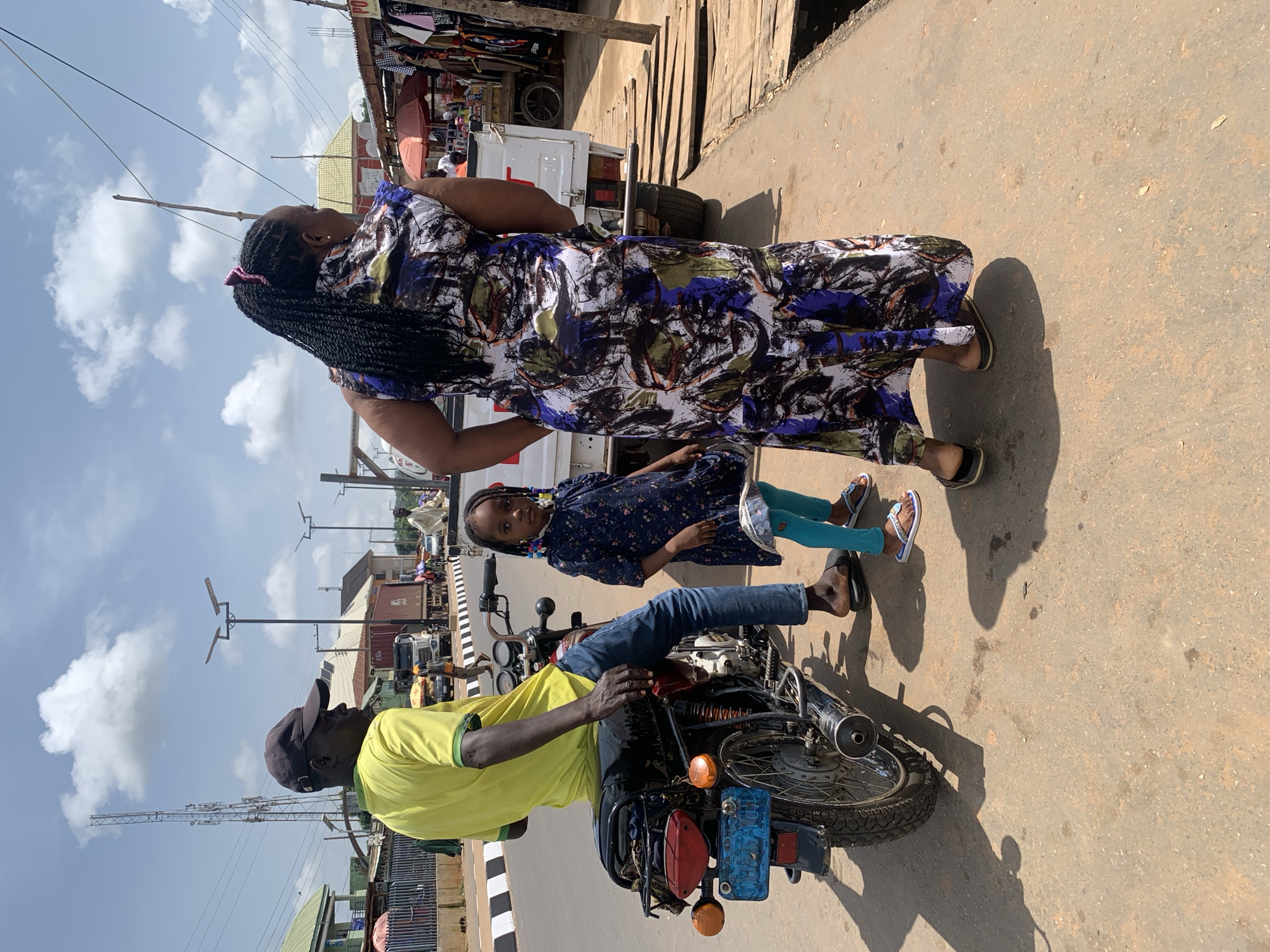 A family is gathered in a street market scene in Akwanga, Nigeria. A woman in a long, patterned dress stands, her back to the camera, with her arm on the shoulder of a young girl. The girl, wearing a dark floral dress and bright turquoise leggings, stands beside her, looking towards the camera. A man, wearing a yellow t-shirt and a dark baseball cap, sits on a motorcycle in the foreground, his hands on the handlebars. The motorcycle is facing to the right, with its rear tire closest to the viewer. Behind the people, market stalls are visible, with various goods displayed. A white truck is parked in the background. The sky is partly cloudy, and the sun casts long shadows, suggesting it is daytime, likely late morning or early afternoon. The atmosphere appears casual and everyday, with no strong emotional cues evident. No visible text is present in the image.