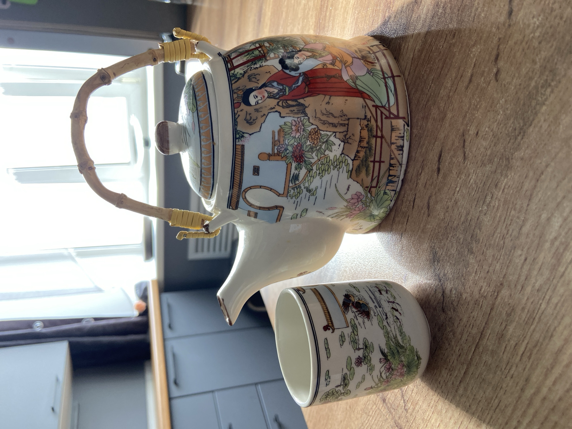 A close-up, eye-level shot shows a white ceramic teapot and cup with traditional East Asian artwork, placed on a wooden table. The teapot is larger, featuring a bamboo handle and a lid, with a scene depicting two women in traditional clothing amidst a garden with water features and flowers. The cup, smaller and to the left of the teapot, has a similar design featuring a body of water with lilies and a duck. The background shows a kitchen with grey cabinets, a wooden countertop, and a window with natural light filtering through. The image is well-lit, with strong shadows cast by the objects. There are no people visible in the image. The scene is static and does not depict any actions or interactions. This image is not located in the city of Tuymazy, Russia.