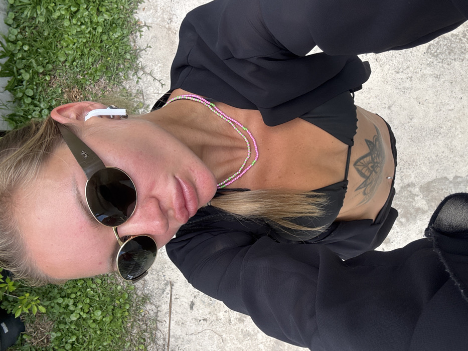 A female individual is depicted in an overhead view, possibly a selfie, against a background of concrete and green foliage in Choeng Thale, Thailand. She has fair skin, light-colored hair, and is wearing dark oval sunglasses with a visible "Ray-Ban" logo on the right temple. A white wireless earbud is present in her right ear. Her lips appear full with a glossy finish. She wears two layered delicate beaded necklaces, one pink and one light green. Her attire includes a black bikini top and a sheer black long-sleeved cover-up, open at the front. A large circular, mandala-style tattoo is visible on her upper abdomen, along with a silver navel piercing. The concrete ground appears rough-textured, and the green foliage consists of small, dense plants.