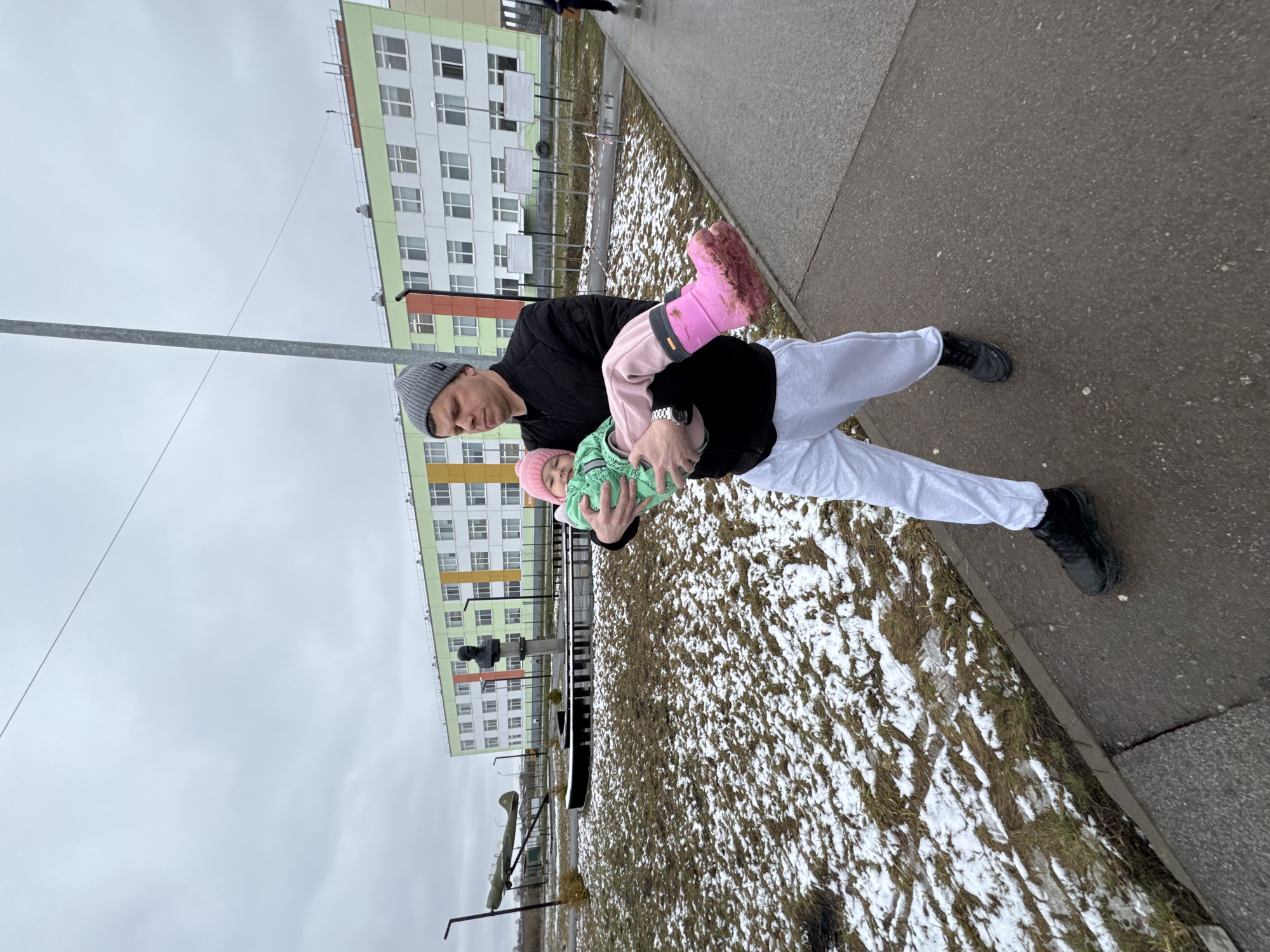 An outdoor image depicts a man carrying a small child in what appears to be a residential area in Горный, Russia. The man, positioned slightly left of center, wears a black jacket, white athletic pants, black shoes, and a grey beanie with a small, unreadable white tag on the front. A watch is visible on his left wrist. He holds a young child, dressed in a light green top and a pink hat, who is nestled against his chest. One of the child's bright pink boots, heavily soiled with mud or dirt, is prominently visible.
The scene suggests cold weather, with patches of melting snow and bare ground intermingling with dried grass. A paved grey sidewalk runs along the right side and foreground, with the snow-dusted grass and dirt patch to the left. In the background, large multi-story apartment buildings with light green and white facades, and some yellow or orange accents, line the street. The sky is overcast and grey, indicating a dull day, possibly late winter or early spring. A thin metal pole, likely a utility or street light pole, spans across the upper part of the image, with fine wires extending from it. The overall impression is that of everyday life in a post-Soviet urban environment during a chilly season.