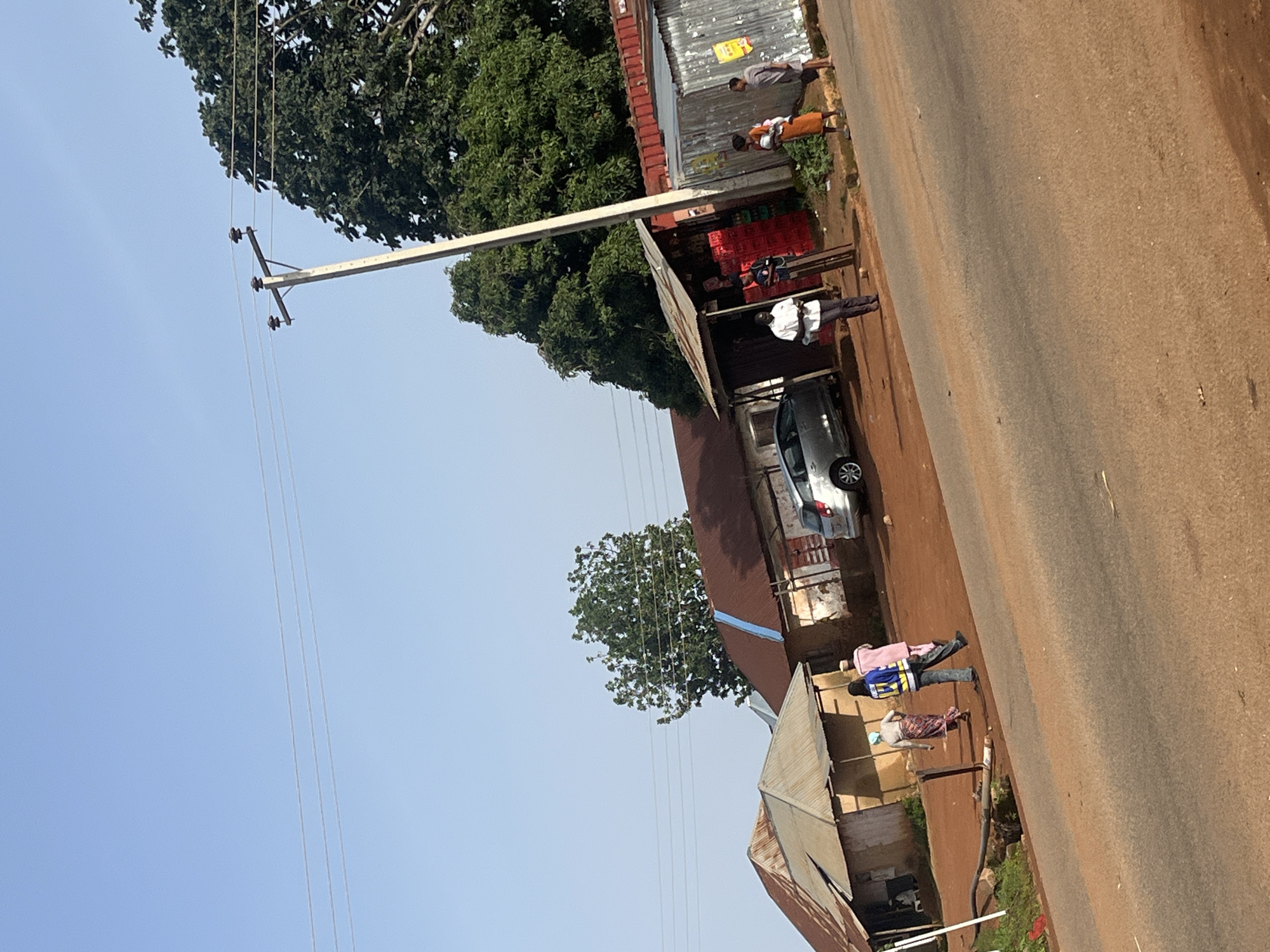 In Kassa, Nigeria, a dirt road runs alongside a few small shops and residential structures. The shops appear to be informal kiosks with corrugated metal roofs and open fronts. One shop displays red crates filled with goods. Several people are visible on or near the road. Two individuals, one in a pink garment and the other in a patterned skirt and headscarf, walk away from the camera on the dirt path. Another person, wearing a blue and yellow reflective vest over dark pants, walks alongside them. Further down the road, three more people stand outside a shop. One person, wearing an orange dress, holds a baby. A silver car is parked in front of one of the structures. A utility pole with wires stretches across the scene, with a large tree providing shade in the background. The sky is clear and blue, suggesting a sunny day. The overall impression is of a busy, albeit modest, commercial and residential area in a Nigerian town.