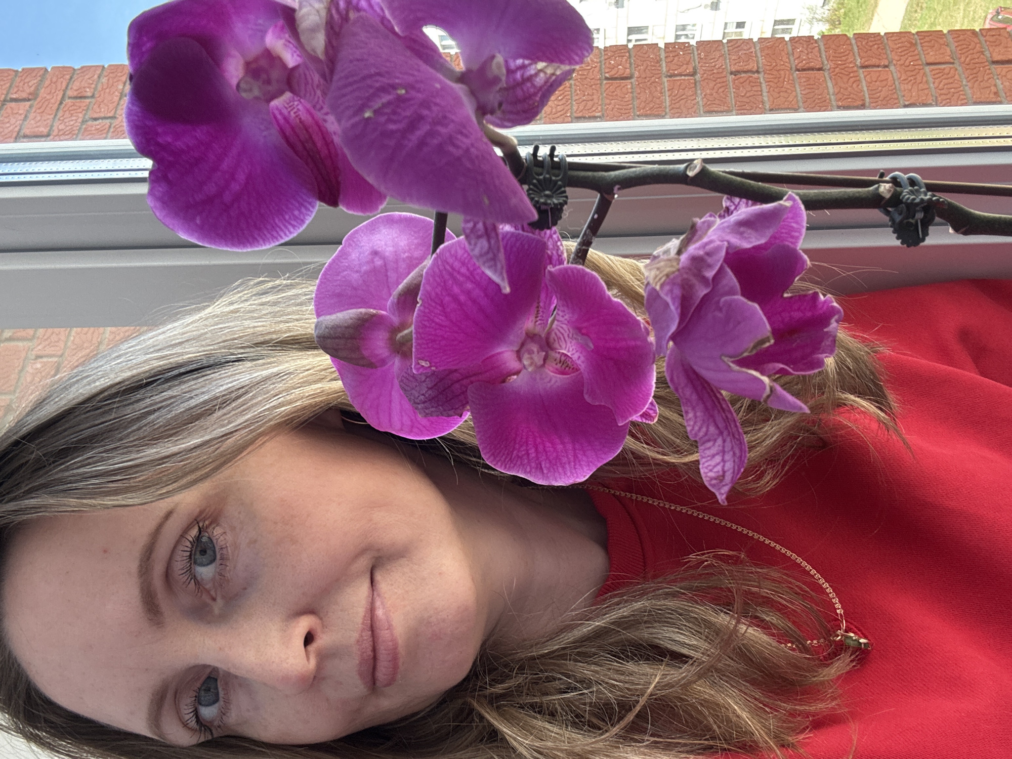 A fair-skinned woman with light-colored hair poses with a spray of pink orchids in front of a window. The woman is wearing a red sweater and a gold chain necklace. Outside the window, a brick building and green space can be seen. The city is Chaikovsky, Russia.