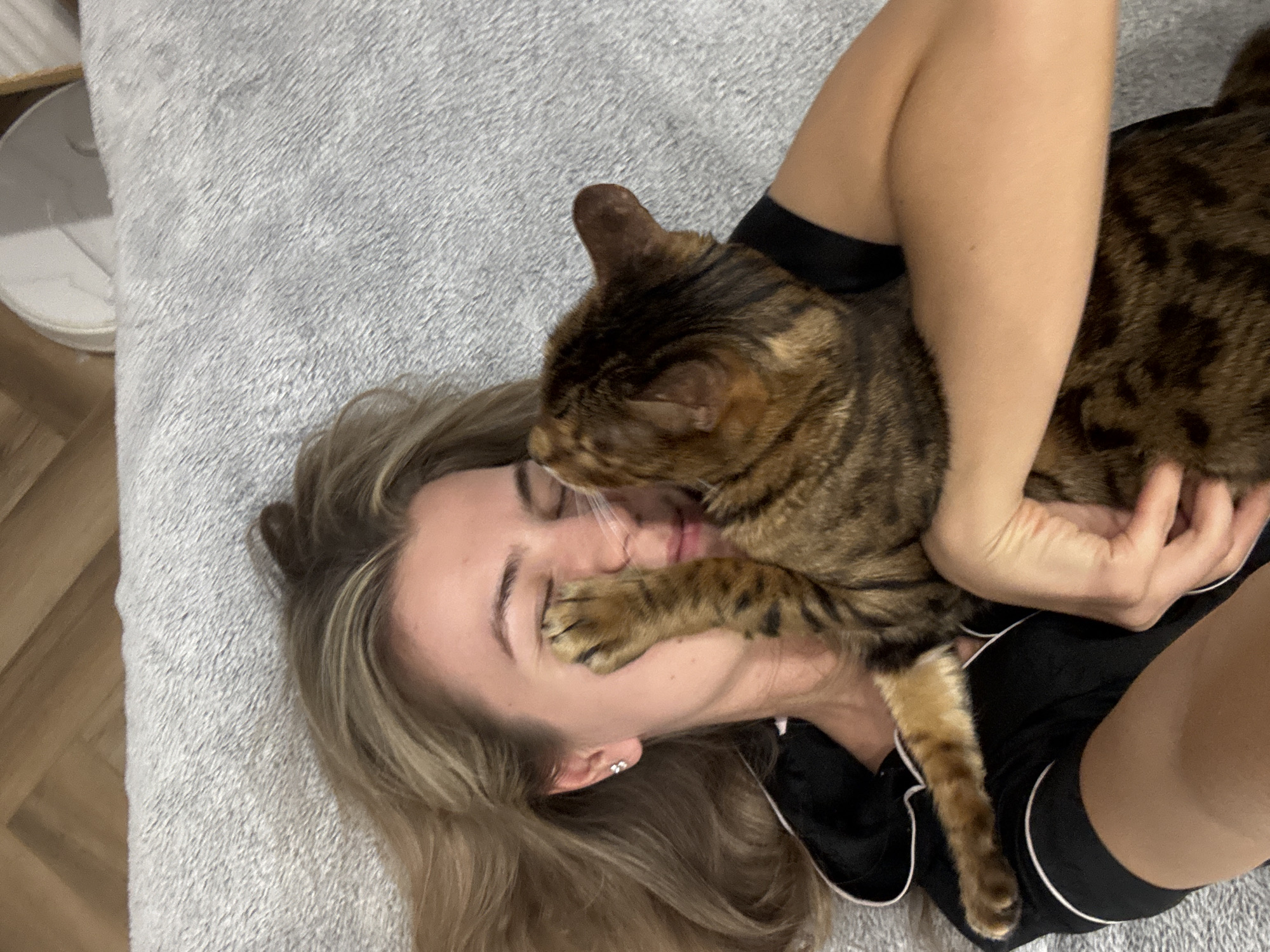 A high-angle, close-up shot shows a woman lying on a gray textured surface with a Bengal cat on her face. The woman is fair-skinned, with blonde hair, and is wearing a black top with white trim. Her right hand is partially visible, holding the cat. The Bengal cat is predominantly brown with darker spots and stripes. It is positioned on the woman's face, its paw resting on her cheek. A round white object is visible in the background near the upper left corner. The scene takes place in an indoor setting.