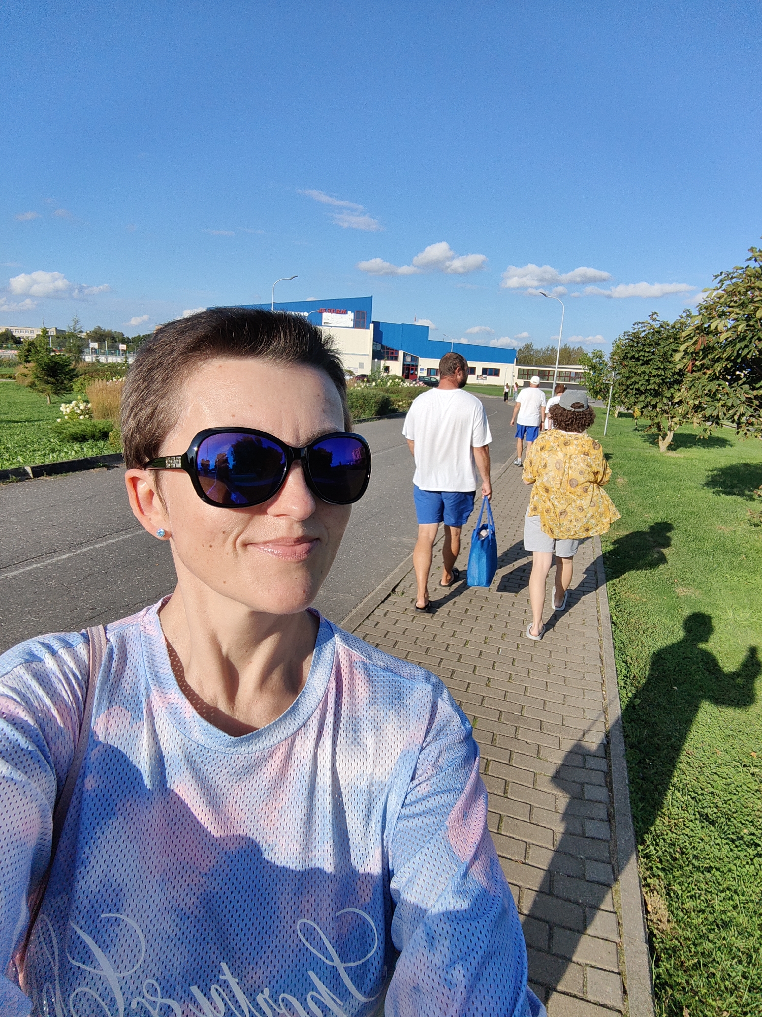 A woman with short, dark hair and sunglasses takes a selfie outdoors. She is wearing a blue and pink tie-dye t-shirt. Behind her, three people walk away on a brick path beside a grassy area. The person closest to the camera is a man wearing a white t-shirt and blue shorts, carrying a blue bag. The second person is wearing a white t-shirt and blue shorts, and the third person is wearing a yellow shirt and gray shorts. In the background, there is a long, blue and white building, trees, and a bright blue sky with scattered clouds. The sun casts long shadows on the grass. The location appears to be the city of Myadzyel, Belarus.
