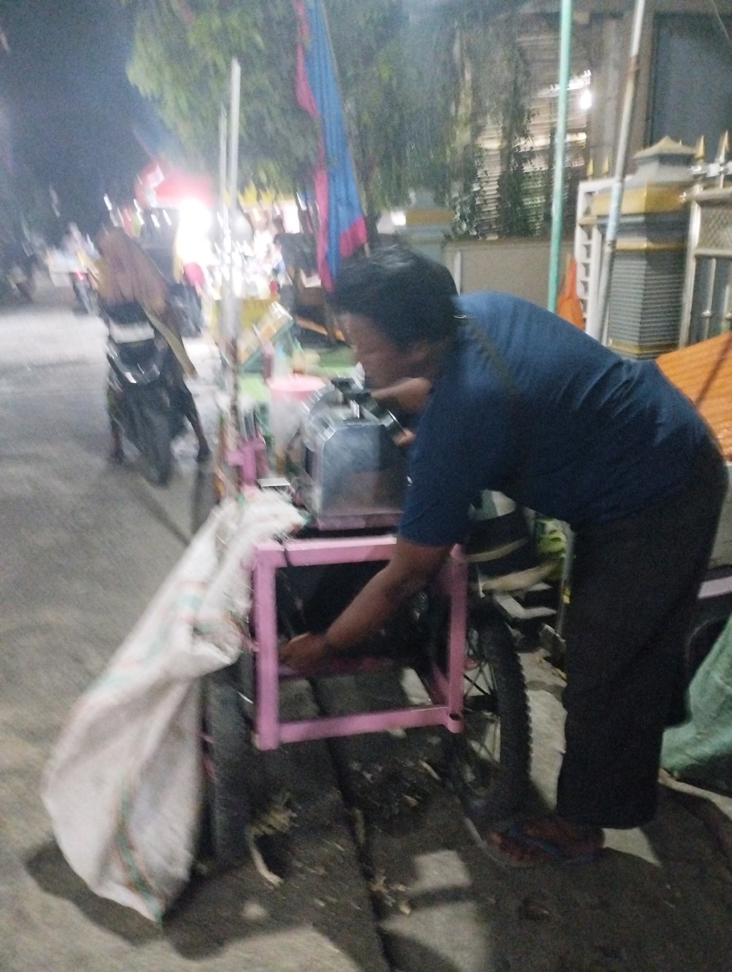 In Jagran Kawistolegi, Indonesia, a person wearing a dark blue t-shirt and black pants is seen leaning over a pink-framed cart. The cart appears to be a mobile vending unit, with a large white sack attached to its side and a metal container on top. The person's hands are engaged with the metal container, suggesting they are operating or attending to it. Behind the cart, a motorcycle is partially visible, and the scene is illuminated by artificial light, indicating it is likely nighttime. The background shows trees, a building with a corrugated metal roof, and street lighting. The ground is paved, and there are other indistinct objects and figures in the periphery, hinting at a busy street or market environment.