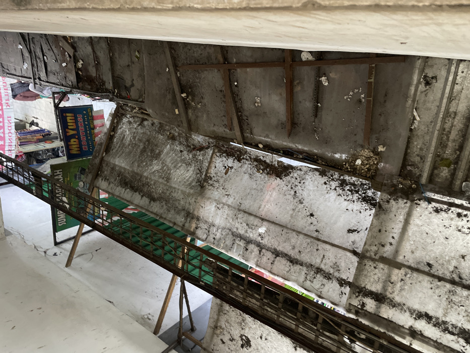 This image provides a high-angle, downward view into a multi-level, neglected commercial passage in Nganguk, Indonesia.

On the left, a dark-colored, vertical metal grate or ladder extends downwards along a faded white wall. Two raw wooden planks are diagonally braced against this wall. The central area is dominated by a translucent, corrugated roof panel, heavily obscured and discolored by widespread dark granular debris and streaks of dirt, which slopes downwards.

To the right, a concrete or tiled inclined surface, possibly a ramp or wide stairwell, descends. This surface is also covered with scattered dark debris and features several irregularly placed horizontal metal or wooden bars. An off-white, textured wall forms the right boundary of the passage.

At the bottom of the frame, indicating a lower commercial level, two signs are clearly visible: a green sign partially obscured, displaying "SUMPAH..." and "MURAH," and a blue sign reading "Mb. Yuni KONVEKSI" with additional text "Pasar Kliwon Blok D" and contact numbers. Below these signs, retail merchandise, including draped fabrics, and a partial human figure in light clothing are faintly discernible. No distinct individuals are present in the immediate foreground or midground. The entire scene indicates significant accumulation of dirt and debris across all surfaces, suggesting a state of disuse or poor maintenance within the commercial structure.